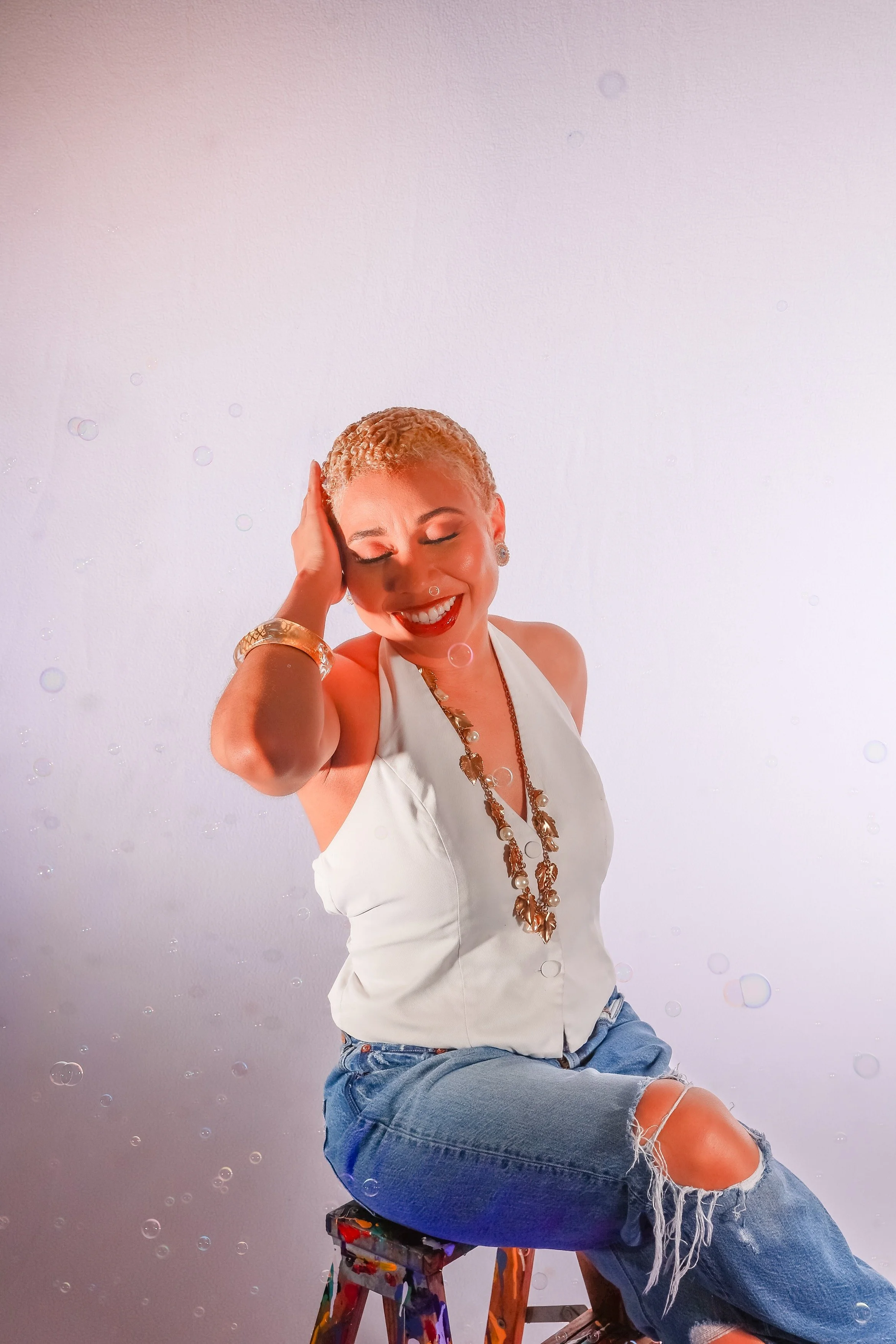 A woman with short blonde hair, white tank top, and ripped jeans sitting on a colorful stool against a plain white wall, smiling with eyes closed and hand on her head.
