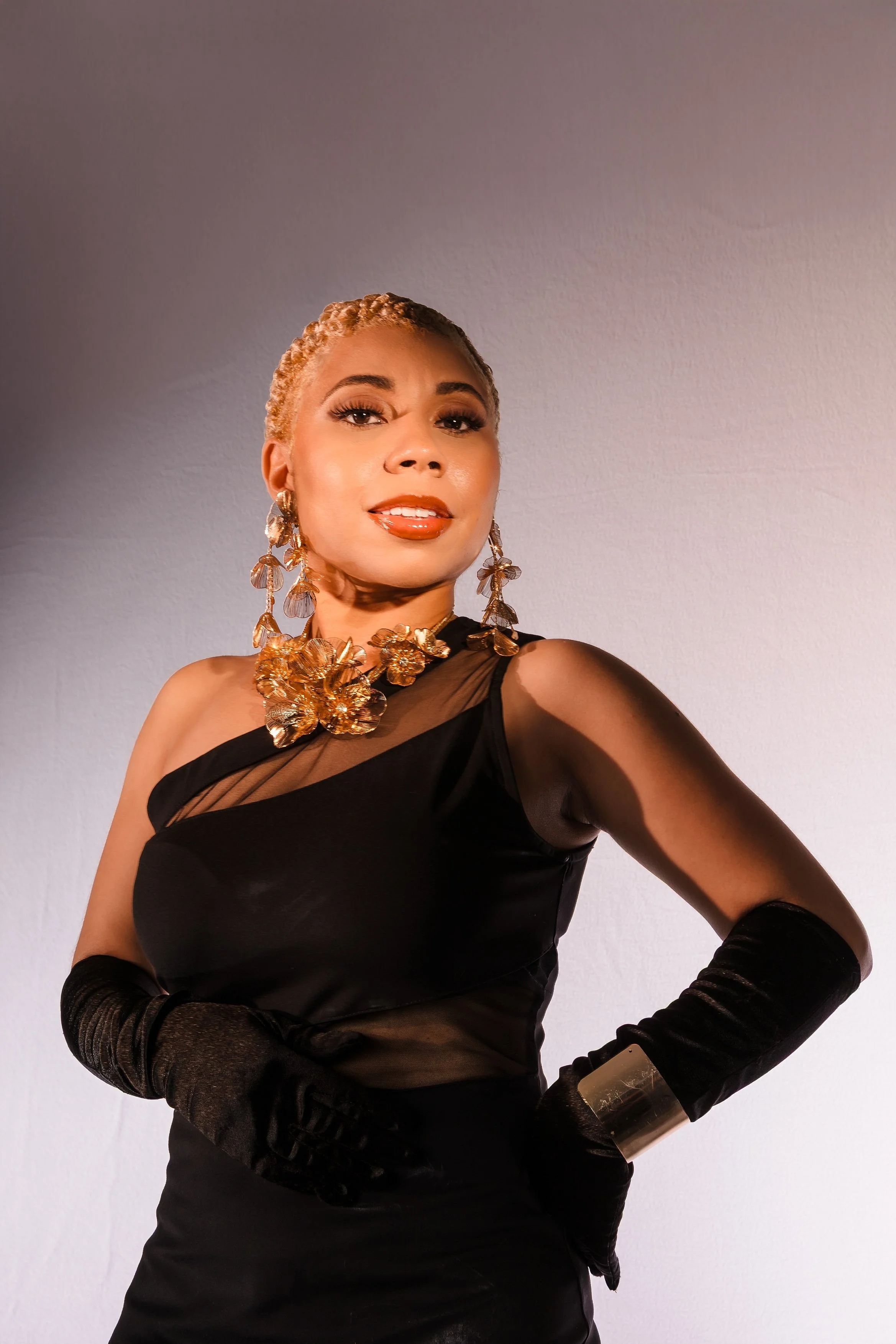 A woman with blonde short hair posing in a black dress with sheer fabric accents, wearing gold jewelry, including large earrings, a statement necklace, and a bracelet, against a plain light-colored background.