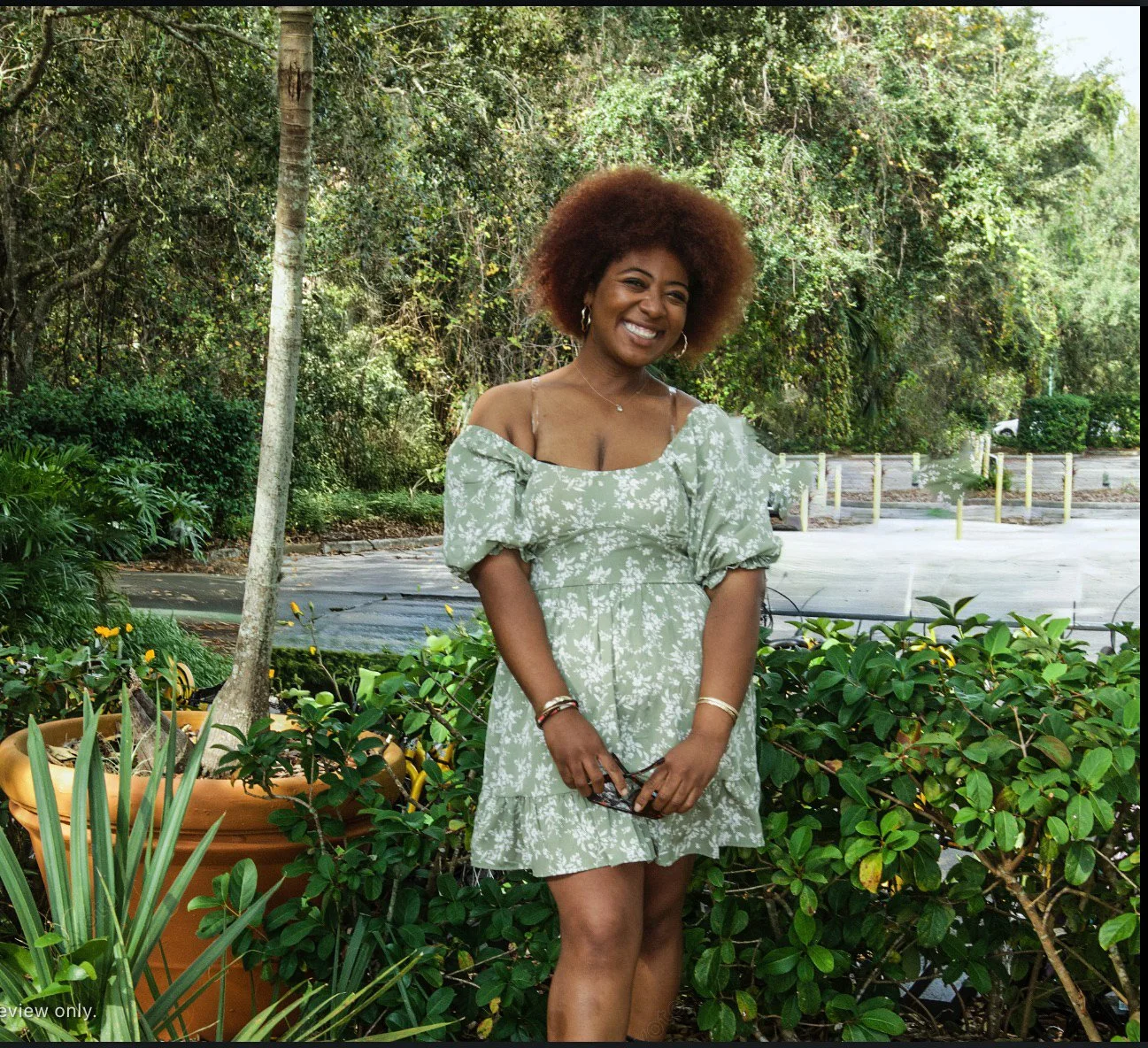 A woman smiling outdoors, wearing a green floral off-the-shoulder dress, standing near green plants and a tree, with trees and a parking lot in the background.