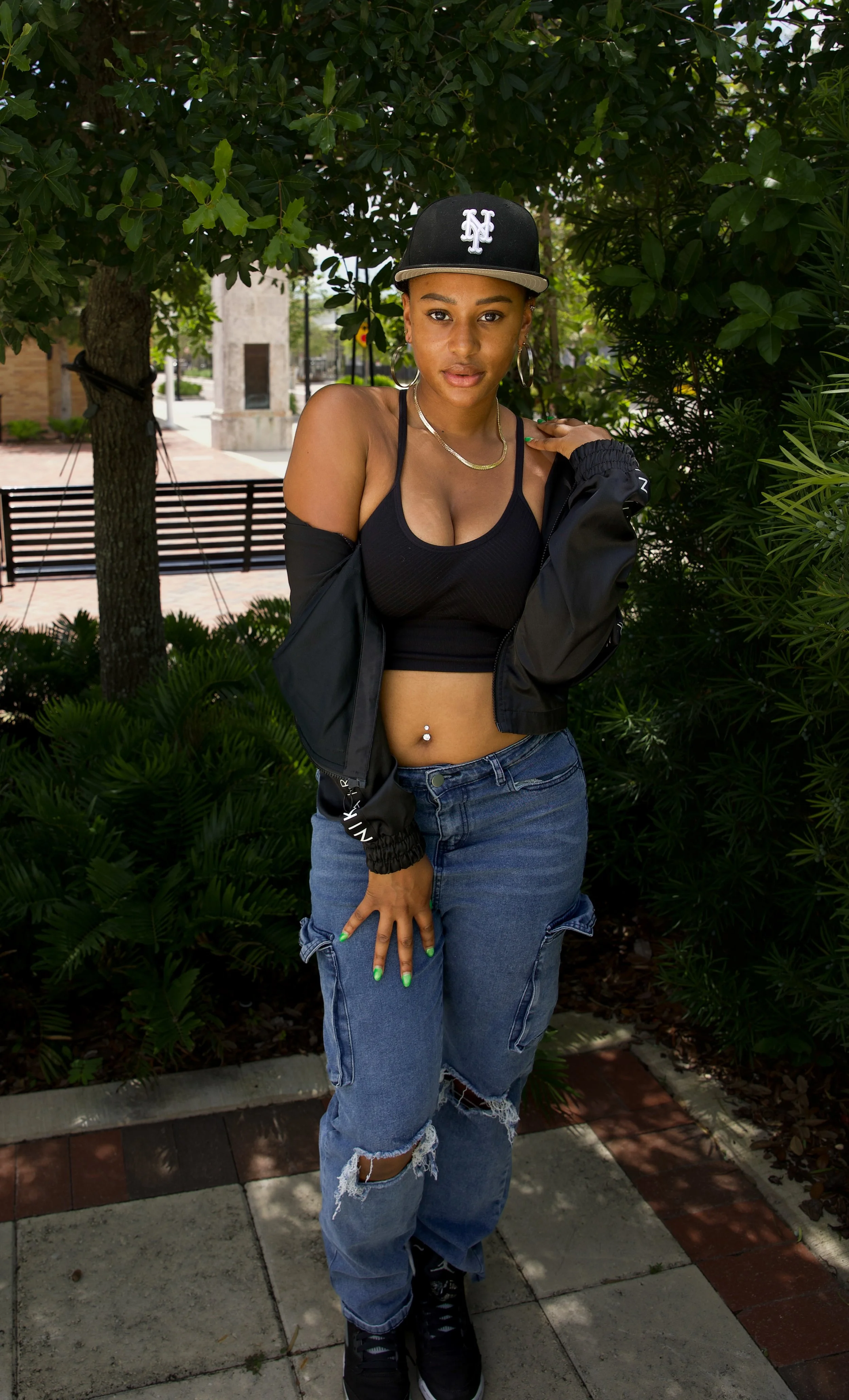 A young woman with medium brown skin and short hair wearing a black baseball cap with a white logo, hoop earrings, layered necklaces, a black crop top, an unzipped black jacket, ripped jeans, and black sneakers, standing outdoors near trees and shrub