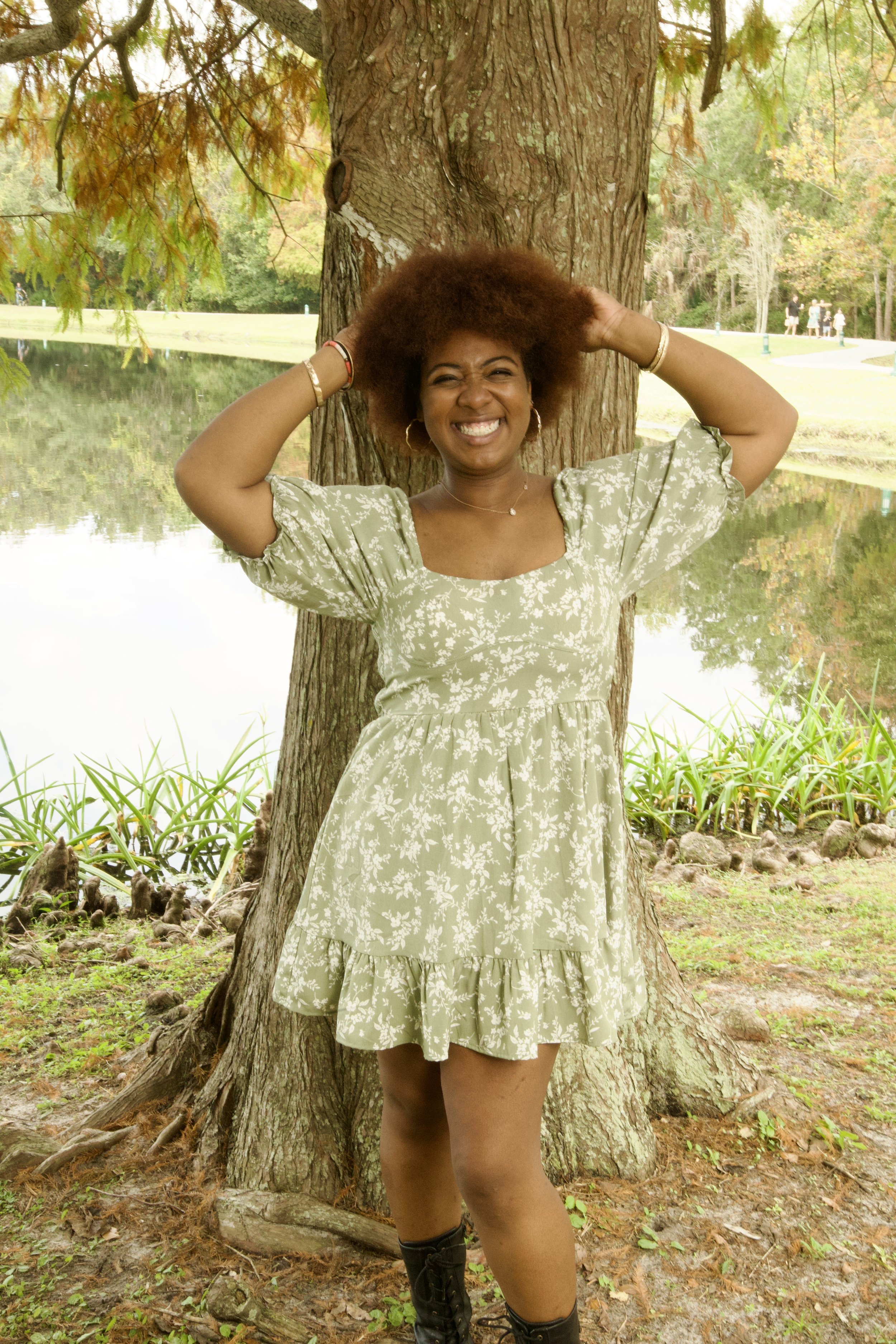 A woman with brown skin and an afro hairstyle, smiling widely, stands outdoors by a tree near a pond. She is wearing a light green floral dress, gold jewelry, and black boots.