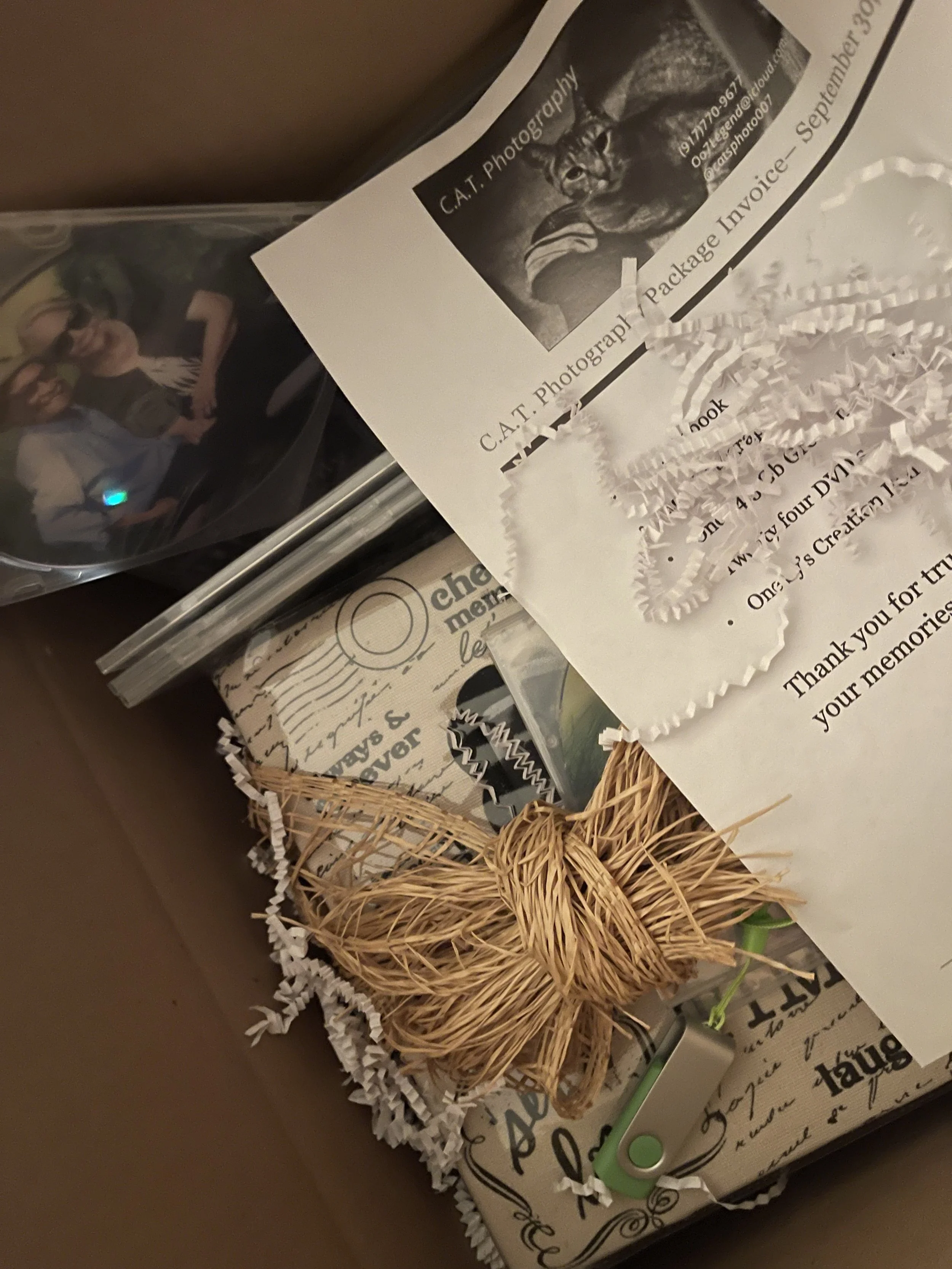 A gift box containing a woven straw butterfly, a photo of two women, a photograph of a cat, printed documents, and a green clip, with white shredded paper for packaging.