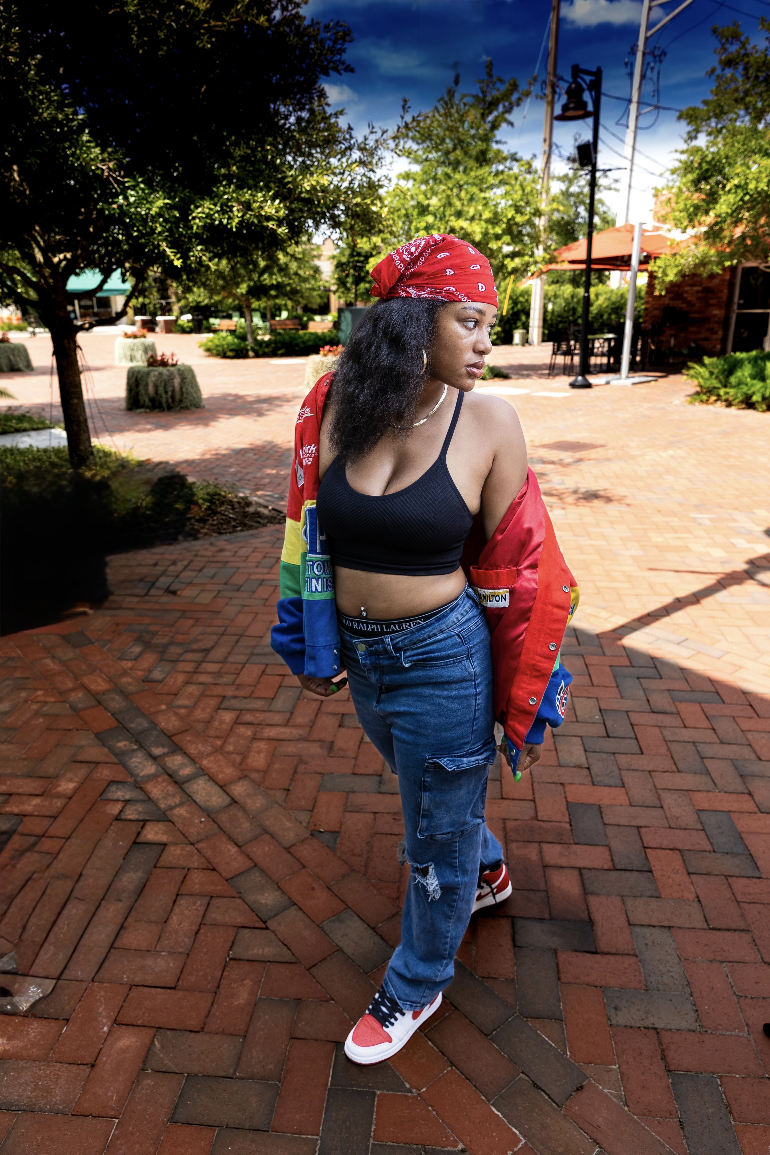 A woman standing outdoors on a brick-paved area under a partly cloudy sky. She is wearing a red bandana, black sports bra, blue jeans, and sneakers, with a colorful jacket draped off her shoulders. She has dark curly hair and is looking to the side.
