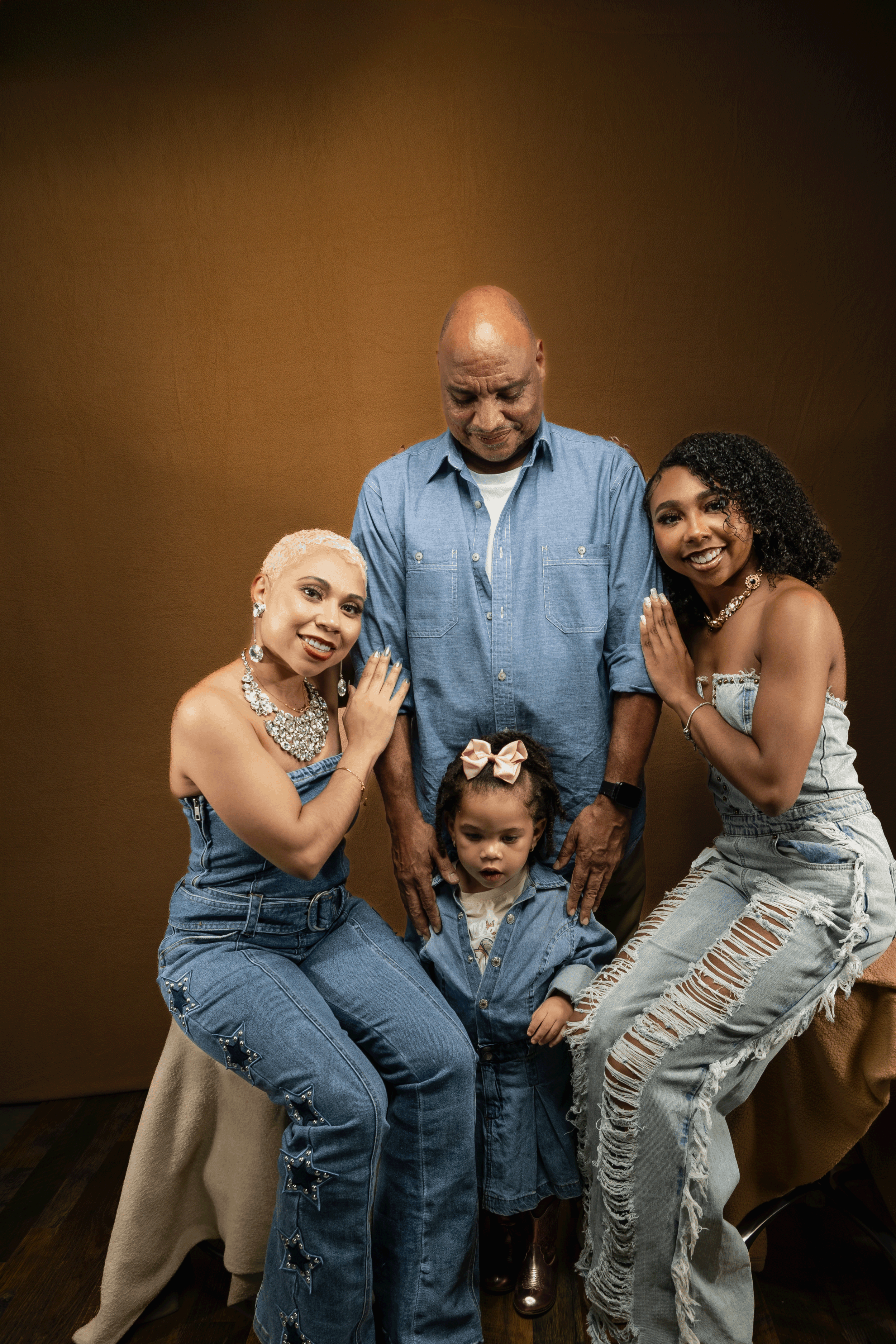 A group of five people, consisting of two adult women, one adult man, and two children, posing together against a brown backdrop. The women are dressed in denim outfits with jewelry, the man is wearing a blue shirt, and the children are also in denim