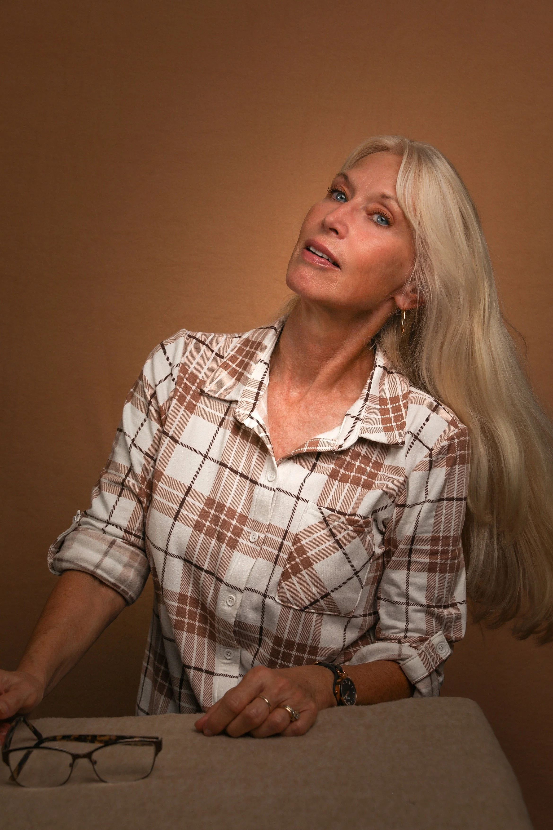 A woman with long blonde hair wearing a plaid shirt with rolled-up sleeves, a watch, rings, and hoop earrings is positioned against a warm brown background. She is looking slightly upward and to her right, leaning over a beige table. A pair of glasses lies on the table in front of her.