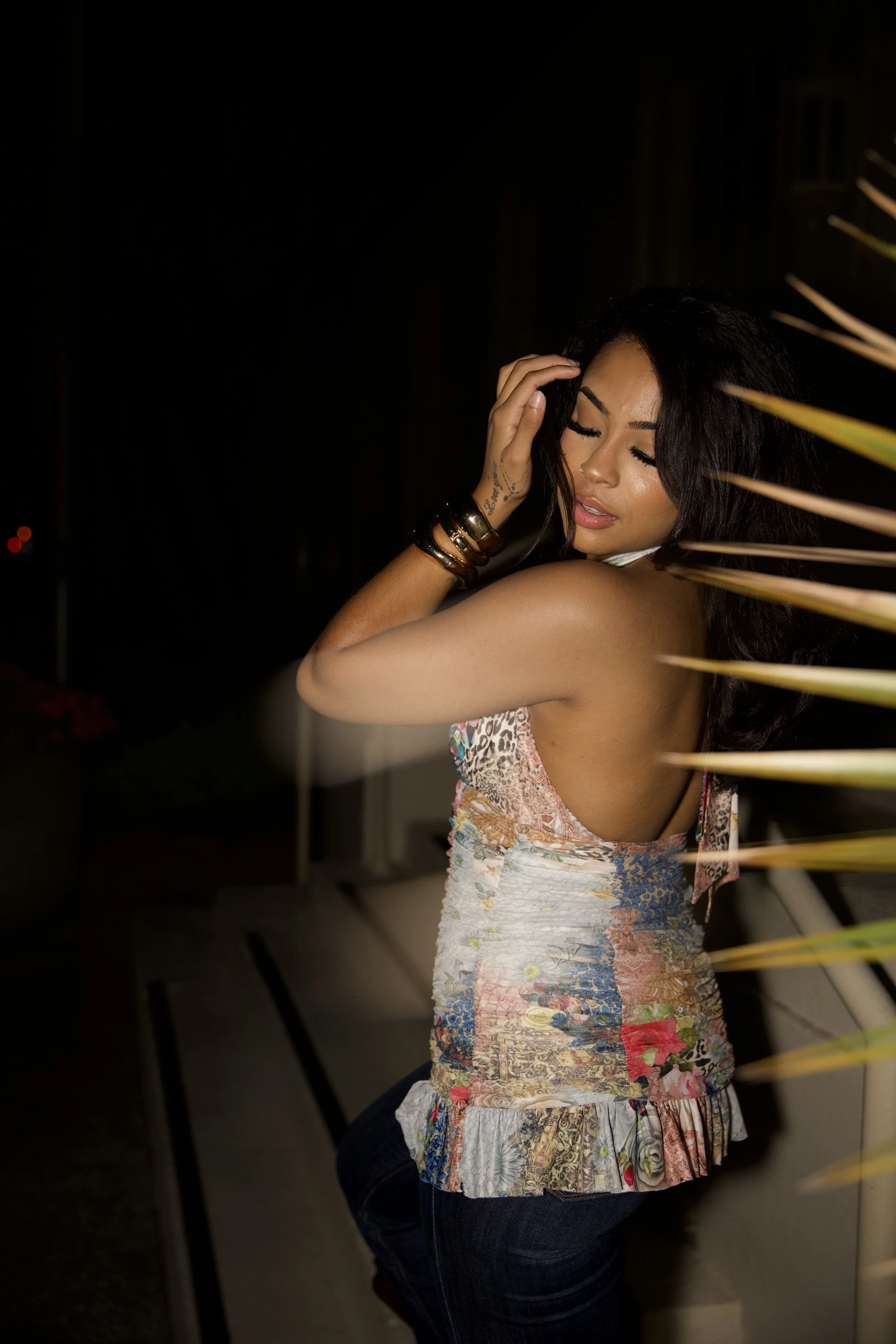 A woman with dark hair and tan skin posing with her eyes closed and one hand on her head, wearing a colorful, patterned sleeveless top and dark jeans, in a dark setting with some illuminated plant leaves in the foreground.