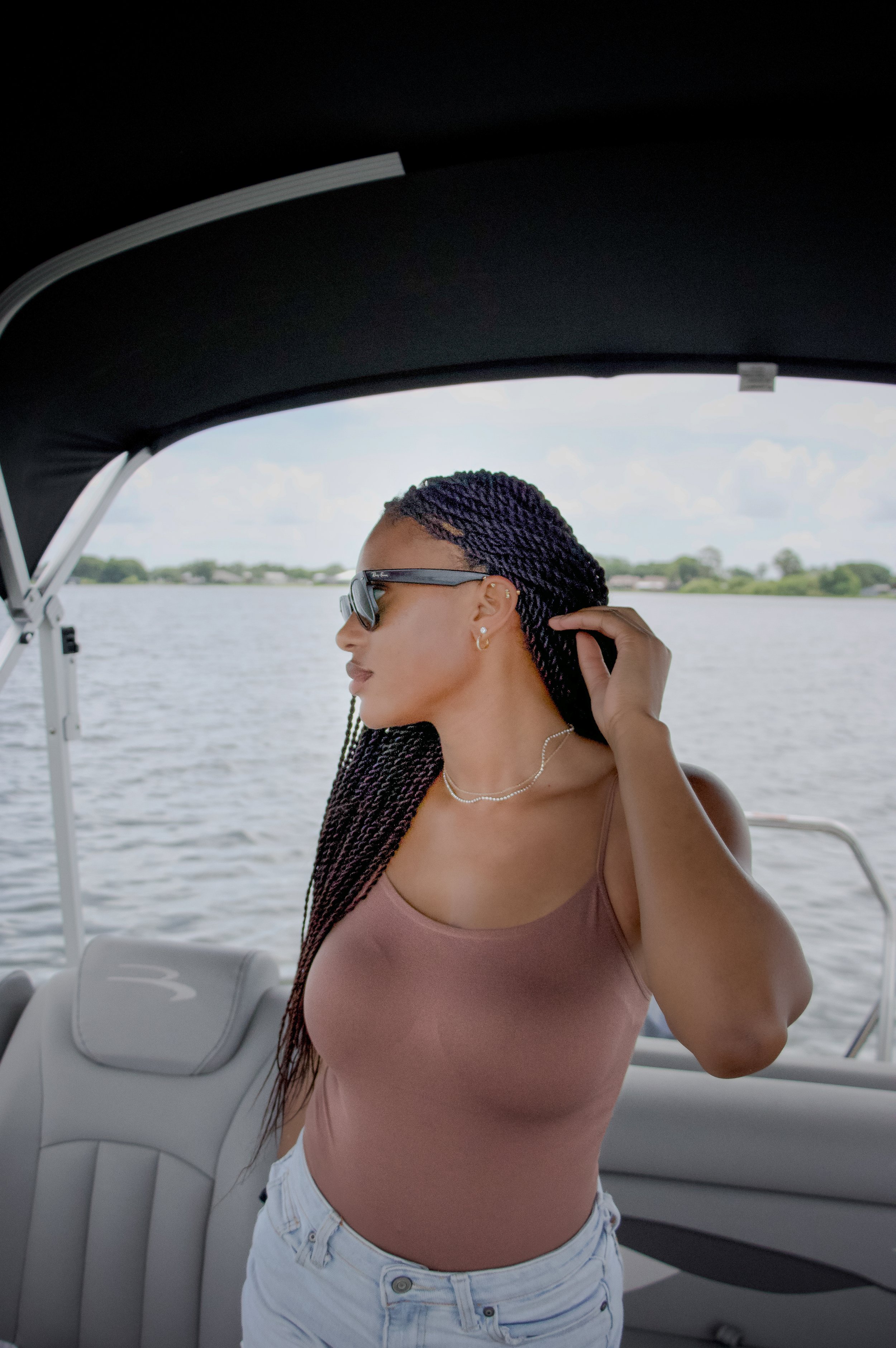 A woman with braided hair wearing sunglasses, a pink tank top, and light-colored jeans on a boat, holding her hair, with water and greenery in the background.