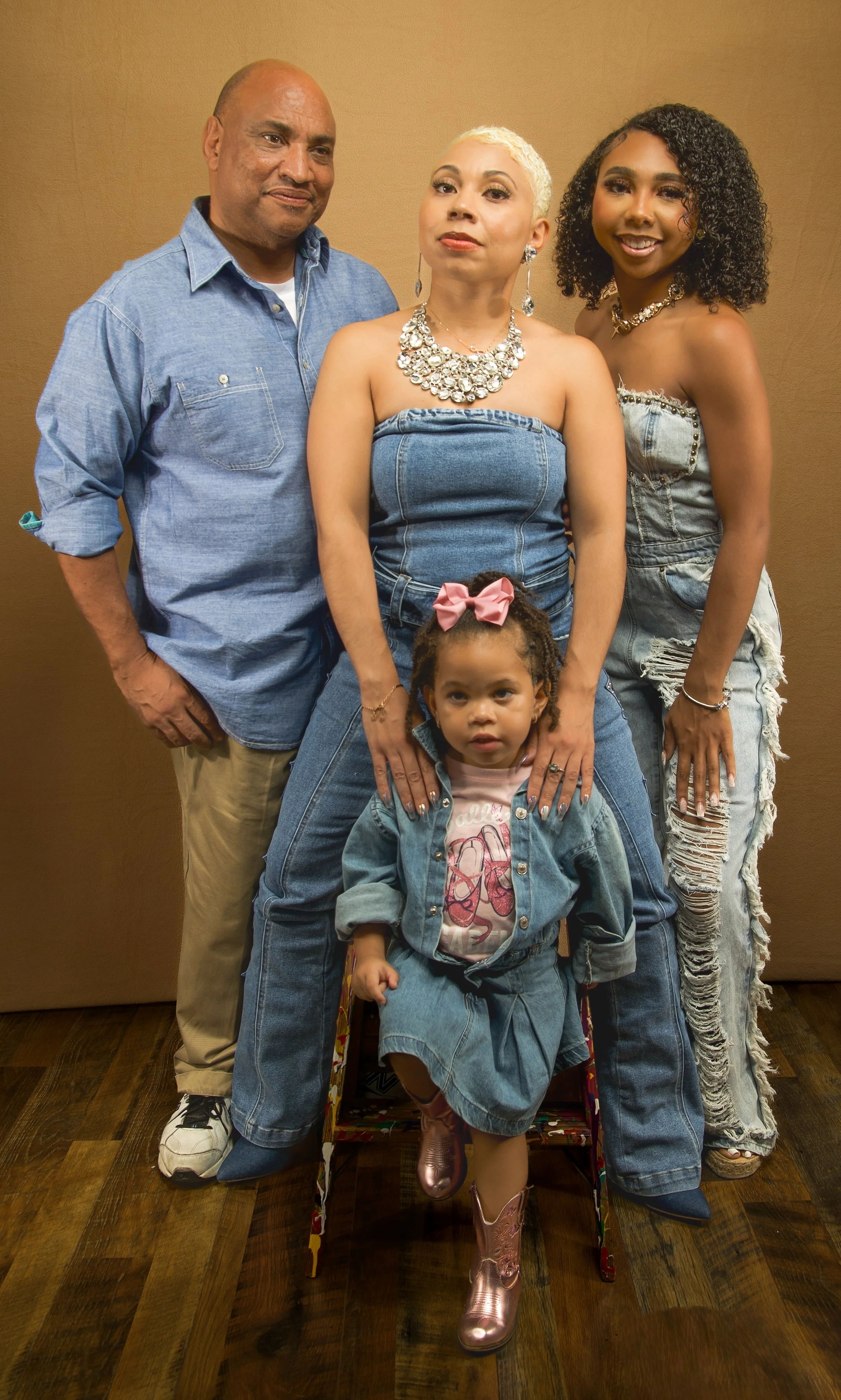 A group of five people, two adults and three children, posing together in front of a brown wall. They are dressed casually, with the woman in the center wearing a denim strapless top and large jewelry, and the children in denim outfits. The group is smiling and looking at the camera.