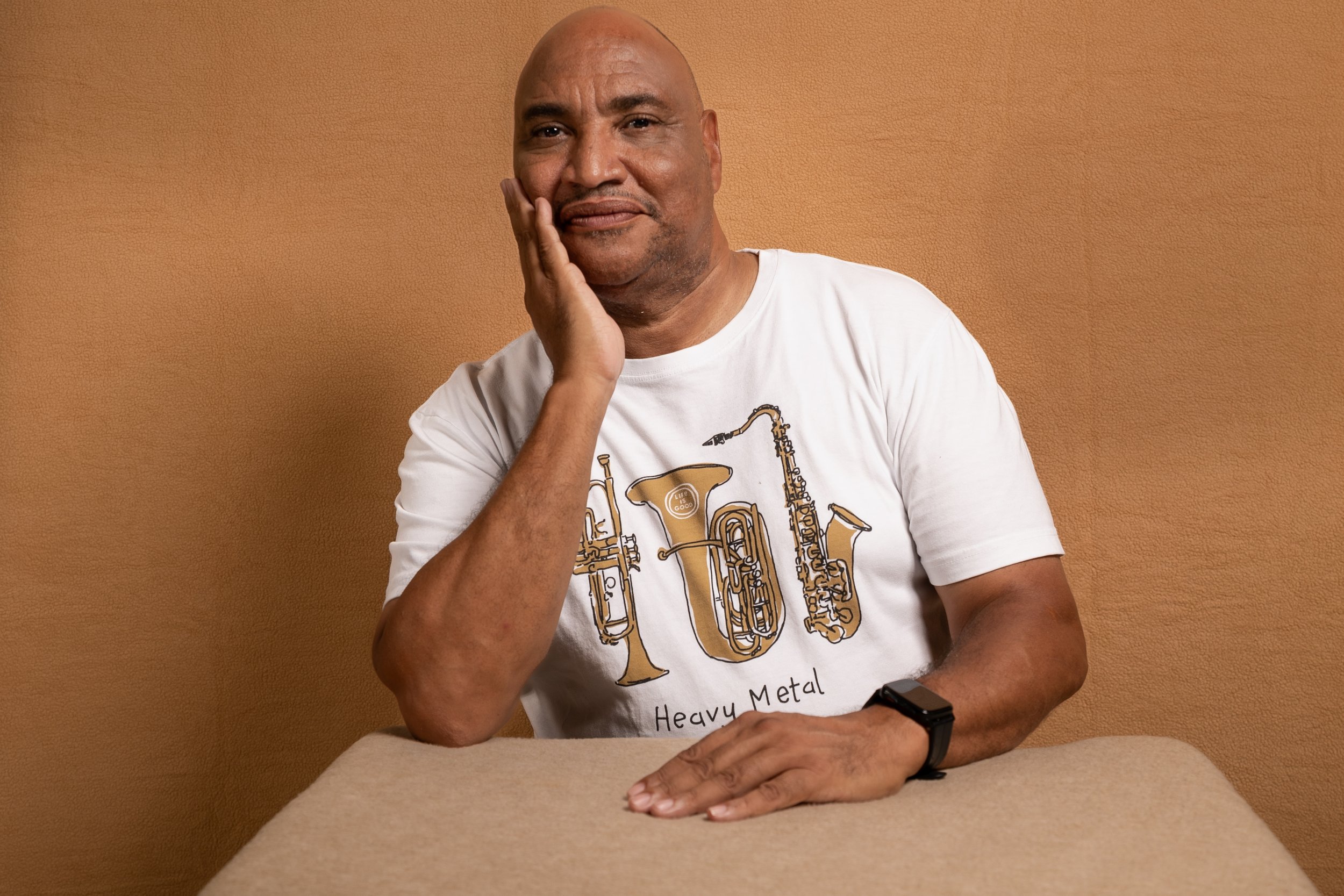 A man with a shaved head and a goatee resting his cheek on his hand, sitting at a table with a beige tablecloth. He is wearing a white T-shirt with a musical instrument design and the words 'Heavy Metal'. The background is a plain brown wall.