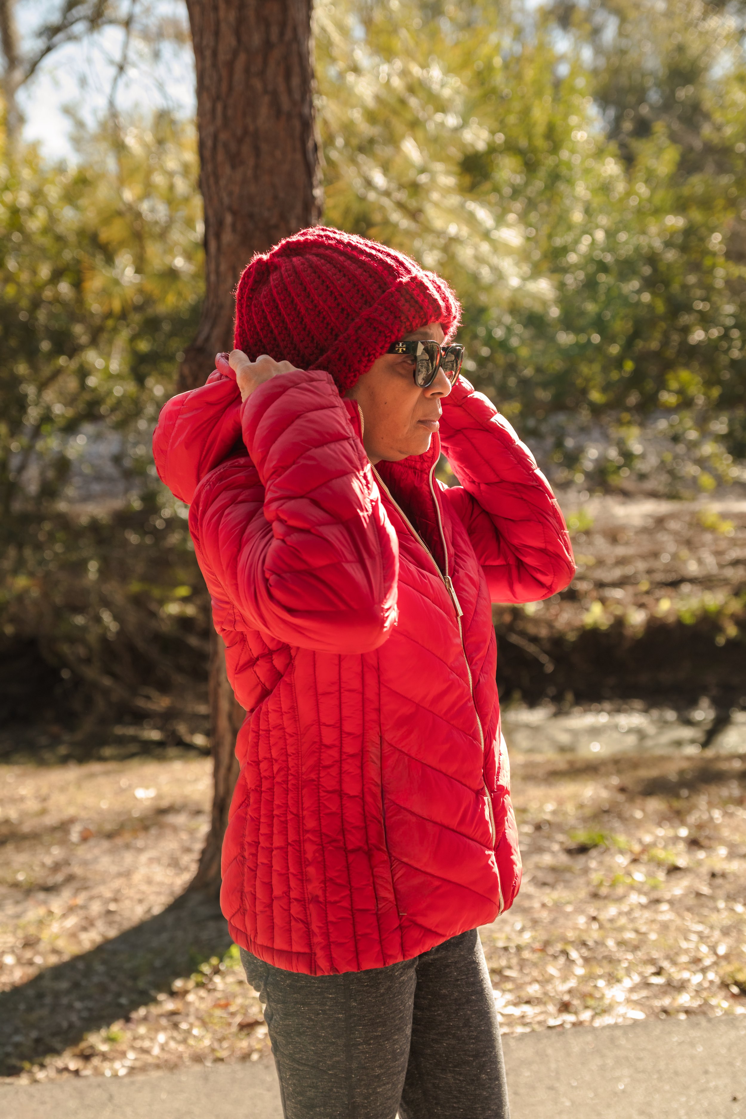 A woman wearing a red knit beanie, sunglasses, a red quilted jacket, and gray leggings standing outdoors near a tree on a sunny day.