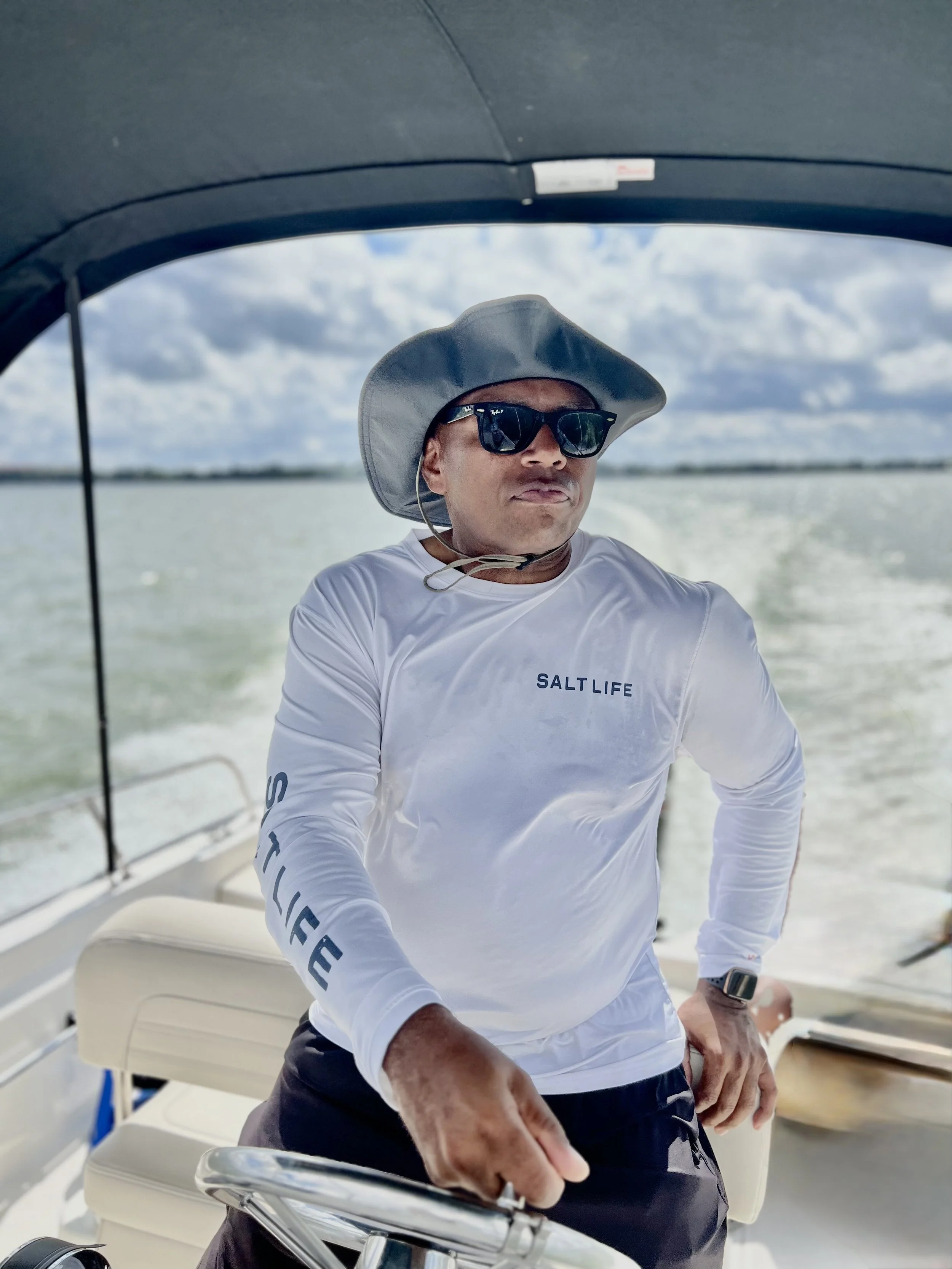 A man wearing a wide-brimmed hat, sunglasses, and a white long-sleeve shirt with 'Salty Life' printed on the front, standing on a boat with a body of water and cloudy sky in the background.