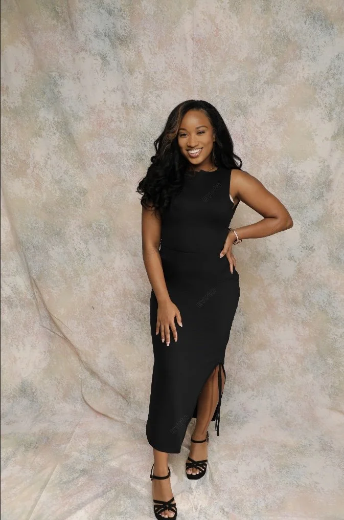A woman in a black sleeveless dress with a slit, wearing black high-heeled sandals, smiling and standing against a textured beige and pink background.