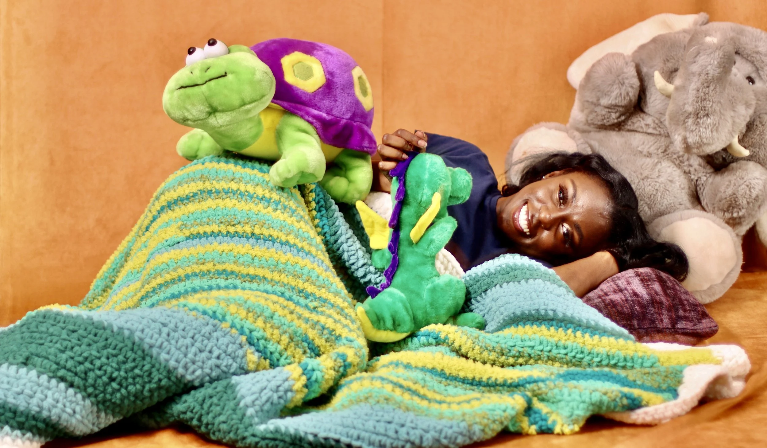 Woman smiling and lying on a bed surrounded by plush toys, including a green turtle with a purple shell and an elephant in the background.