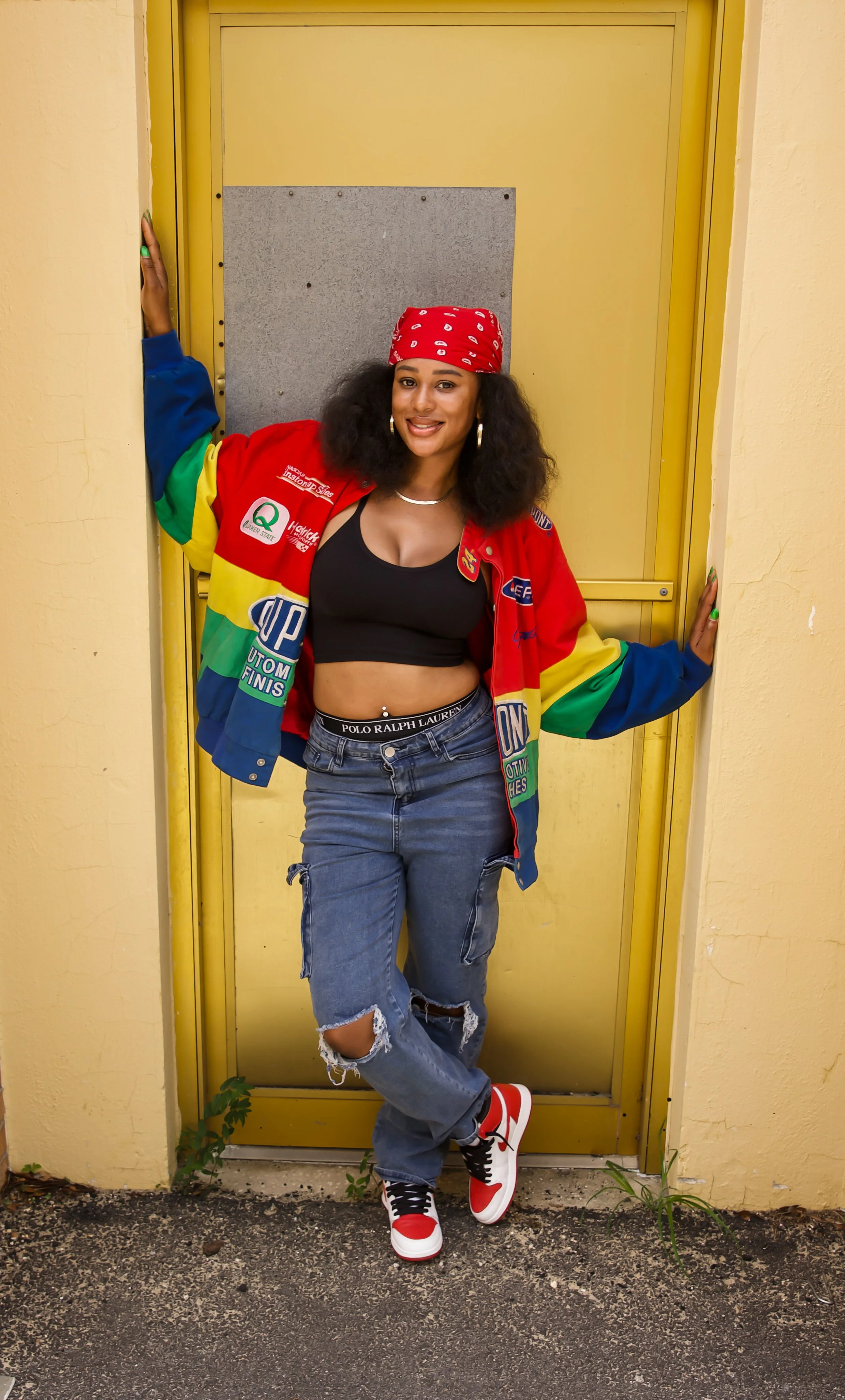A smiling woman with curly black hair, wearing a red bandana, colorful jacket, black crop top, ripped jeans, and red-and-white sneakers, standing in front of a yellow door.