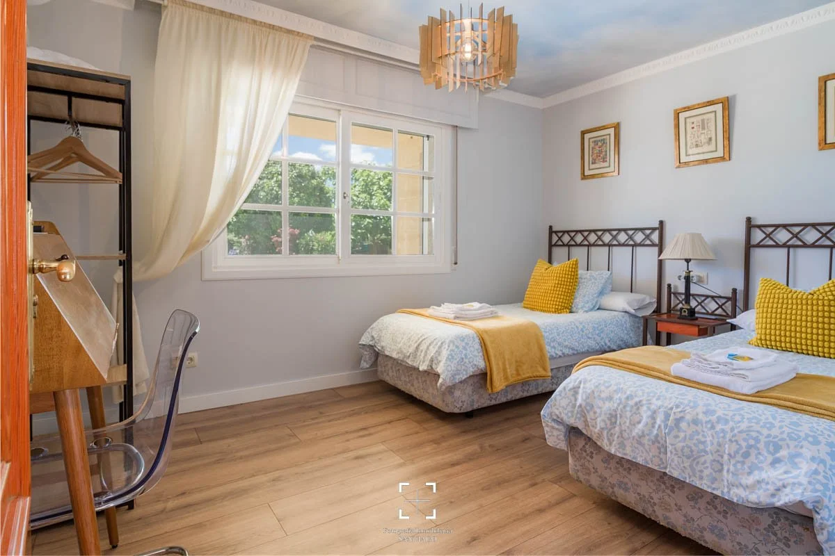 Habitación con dos camas individuales con almohadas amarillas y edredones naranjas, lámpara de mesa, marco de fotos en la pared, ventana grande con cortinas blancas y vista a árboles verdes, suelo de madera, decoración sencilla y luminosa.
