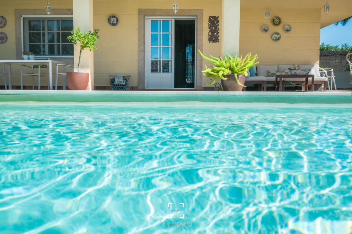 Vista frontal de una piscina con agua cristalina frente a una casa de paredes beige, con una puerta de cristal, plantas en macetas y decoración en la pared exterior, en un día soleado.