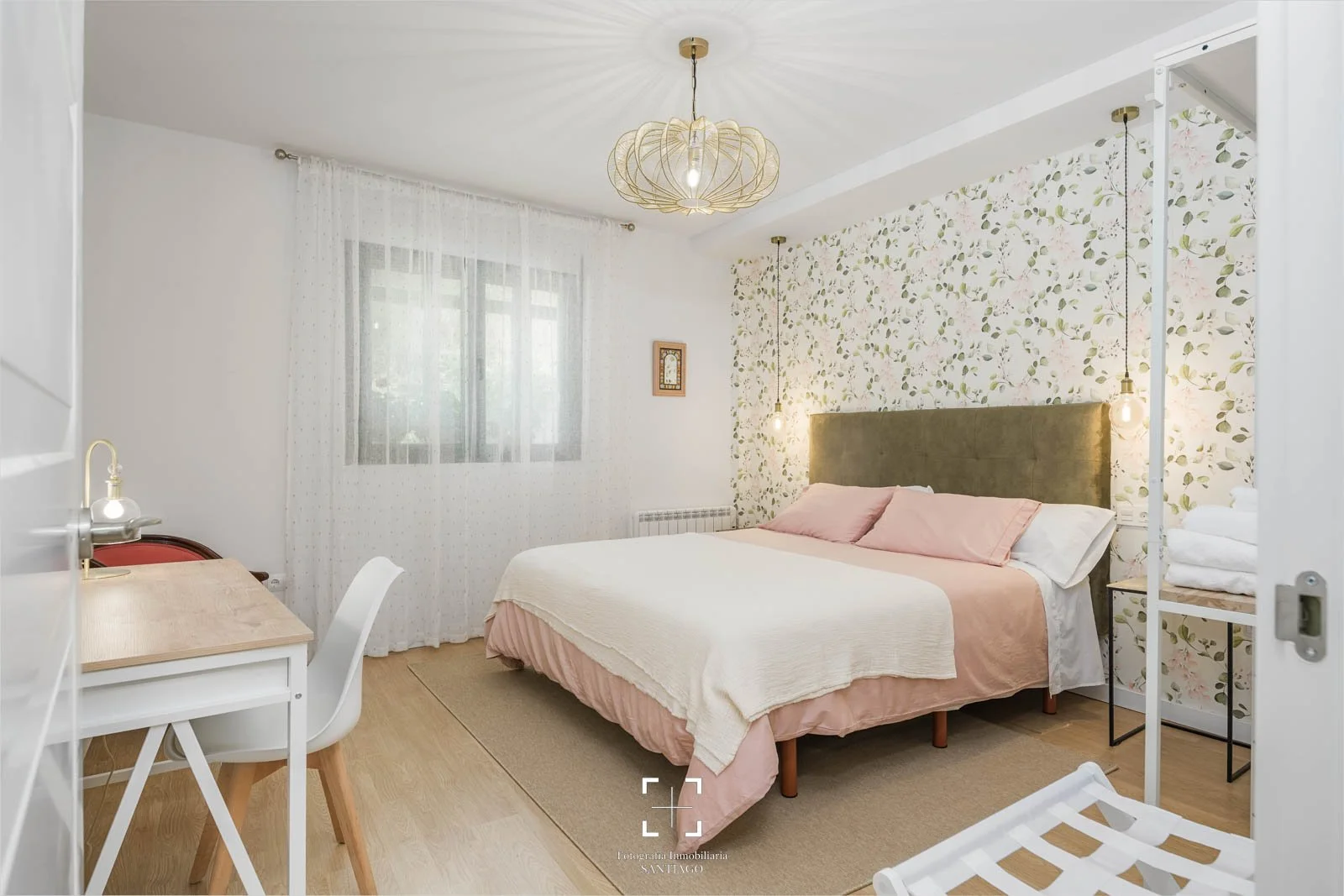 Habitación con cama de matrimonio, paredes decoradas con papel tapiz con hojas, cortinas blancas, lámparas con bombillas expuestas y muebles en tonos claros.