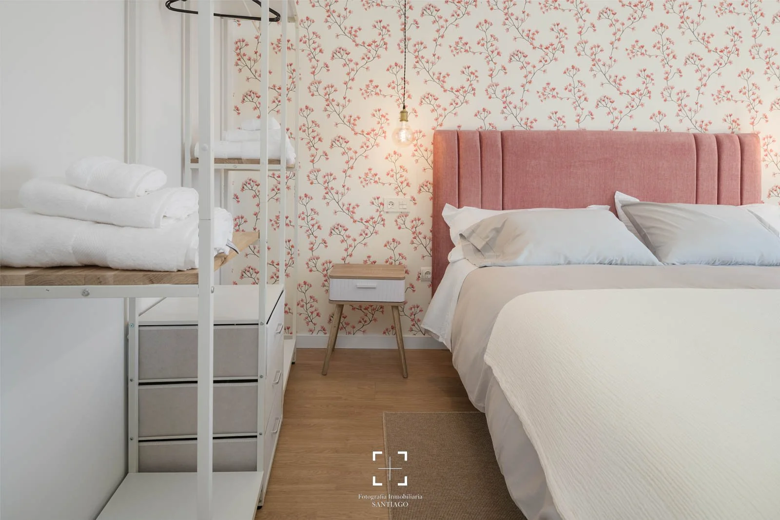 Habitación con paredes de papel tapiz con flores rosadas, cama con cabecera rosa y sábanas blancas, lámpara de bombilla expuesta, mesita de noche de madera, y una litera con ropa blanca.