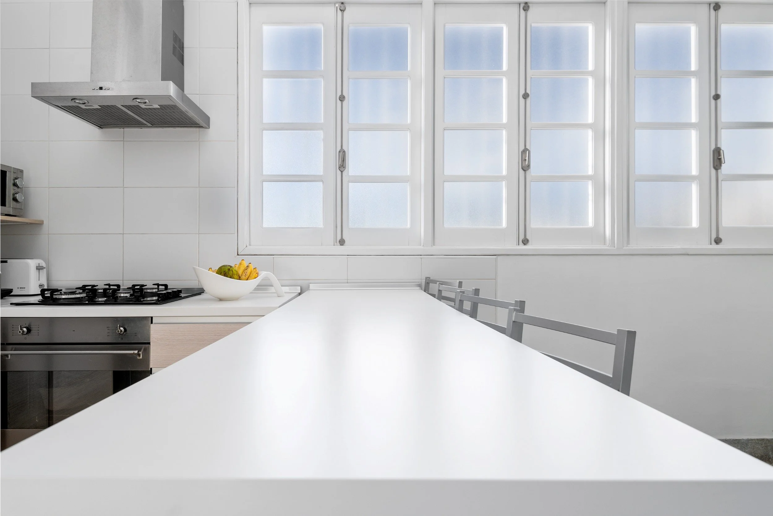 Cocina moderna con mesón blanco, estufa, campana extractora y ventanas grandes con celosías blancas
