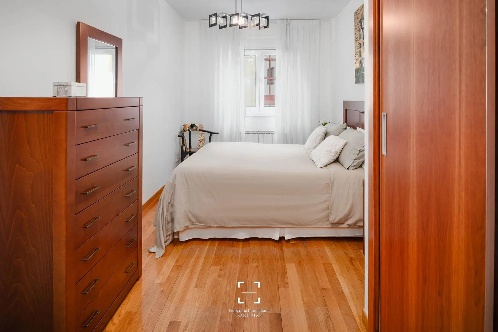 Habitación con cama queen, madera de suelo y muebles, cortinas blancas, ventana, una silla con tapizado y decoración moderna.