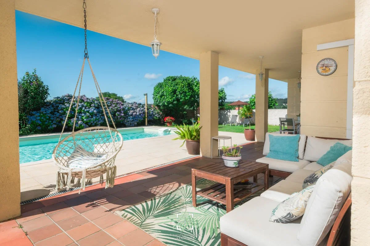 Área de terraza con sillones blancos, mesa de madera, plantas, columpio de mimbre, vista a piscina y jardín con flores y árboles.