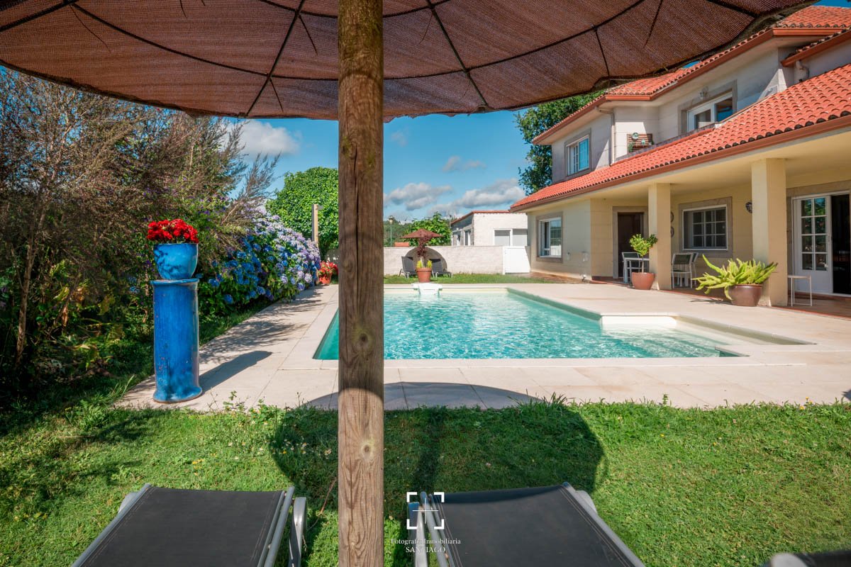 Vista de una piscina en el patio trasero de una casa con plantas y flores, una sombrilla de color marrón, dos sillas de malla y un ambiente soleado con cielo azul