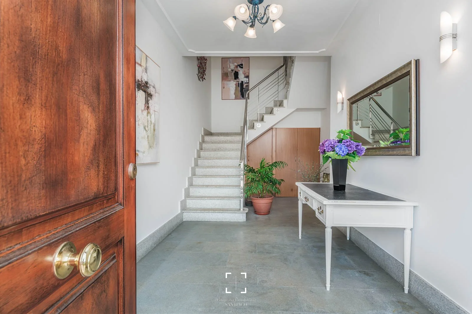 Vista del vestíbulo de una casa, con una puerta de madera a la izquierda, una escalera de mármol blanco en el centro y un espejo grande en la pared derecha reflectando la escalera. Hay una mesa blanca con una planta y un florero con flores moradas so