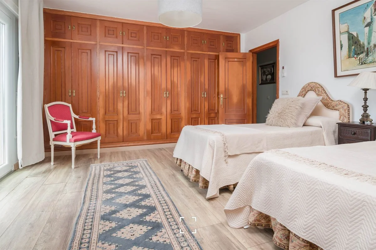 Dos camas con cubrecamas blancos y cojines, una lámpara de mesa, un cuadro en la pared, un armario de madera grande y una silla rosa en la esquina, en una habitación con suelo de madera y cortinas claras.