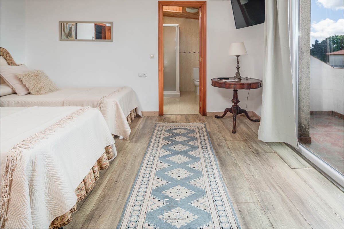 Habitación con dos camas, alfombra azul y beige, mesa antigua con lámpara, televisión en la pared, puerta al baño y balcón con cortinas blancas.