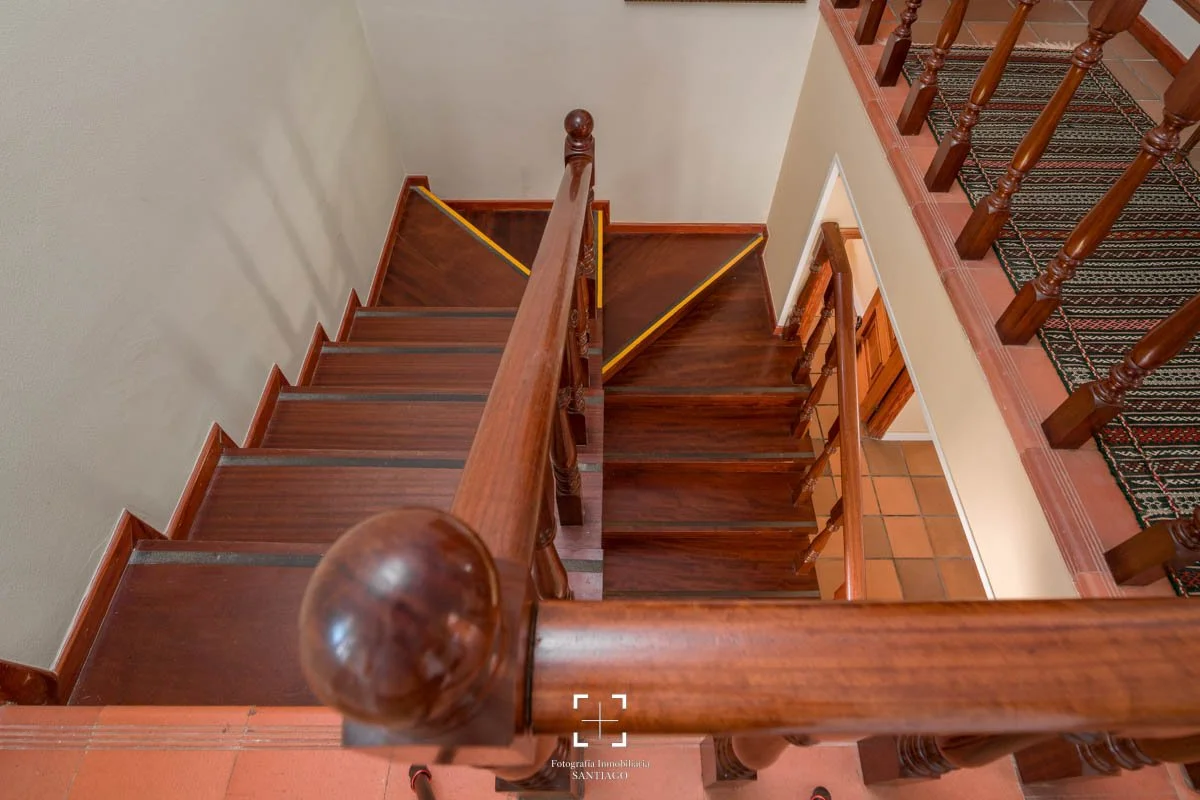 Imagen de una escalera de madera con pasamanos y barandales, vista desde arriba hacia abajo, en una casa. La escalera tiene varias vueltas y un tapete en el piso de la parte superior.