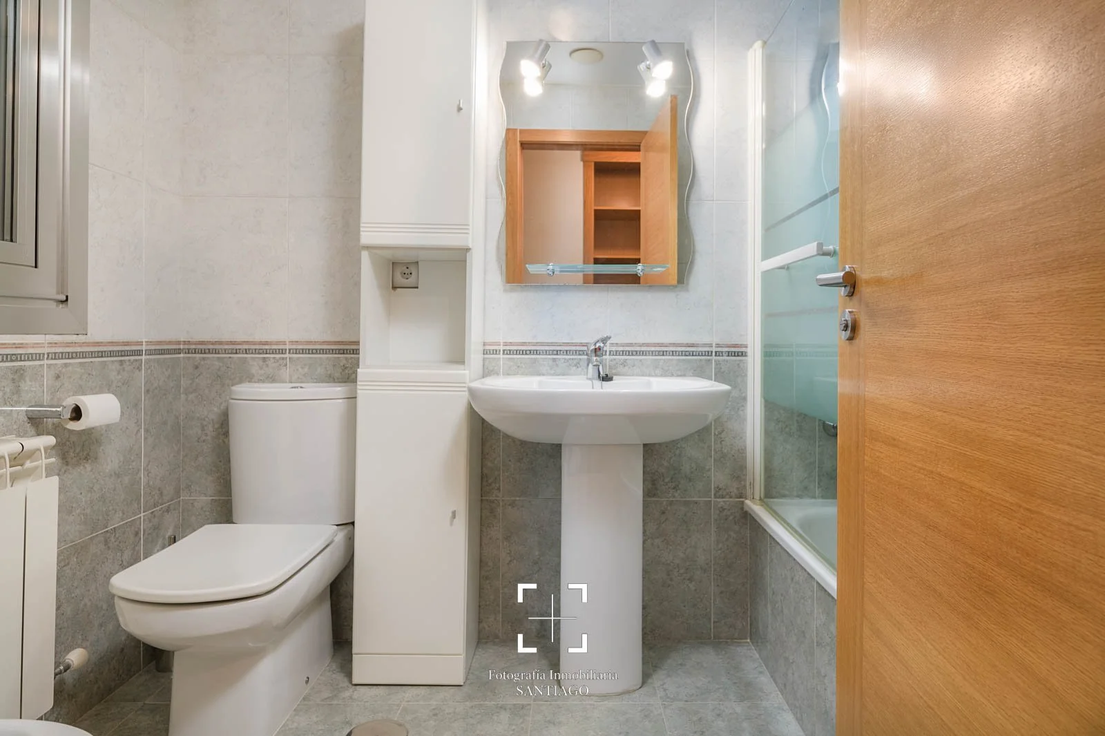 Vista de un baño pequeño con inodoro, lavabo con espejo, ducha y puerta de madera.