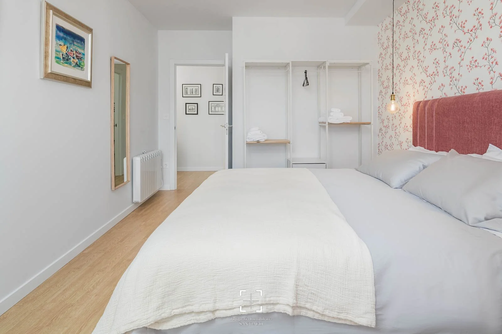 Habitación con cama grande, paredes blancas, pared con papel de pared floral rosa, estantería con toallas, lámpara colgante y obras de arte en las paredes.