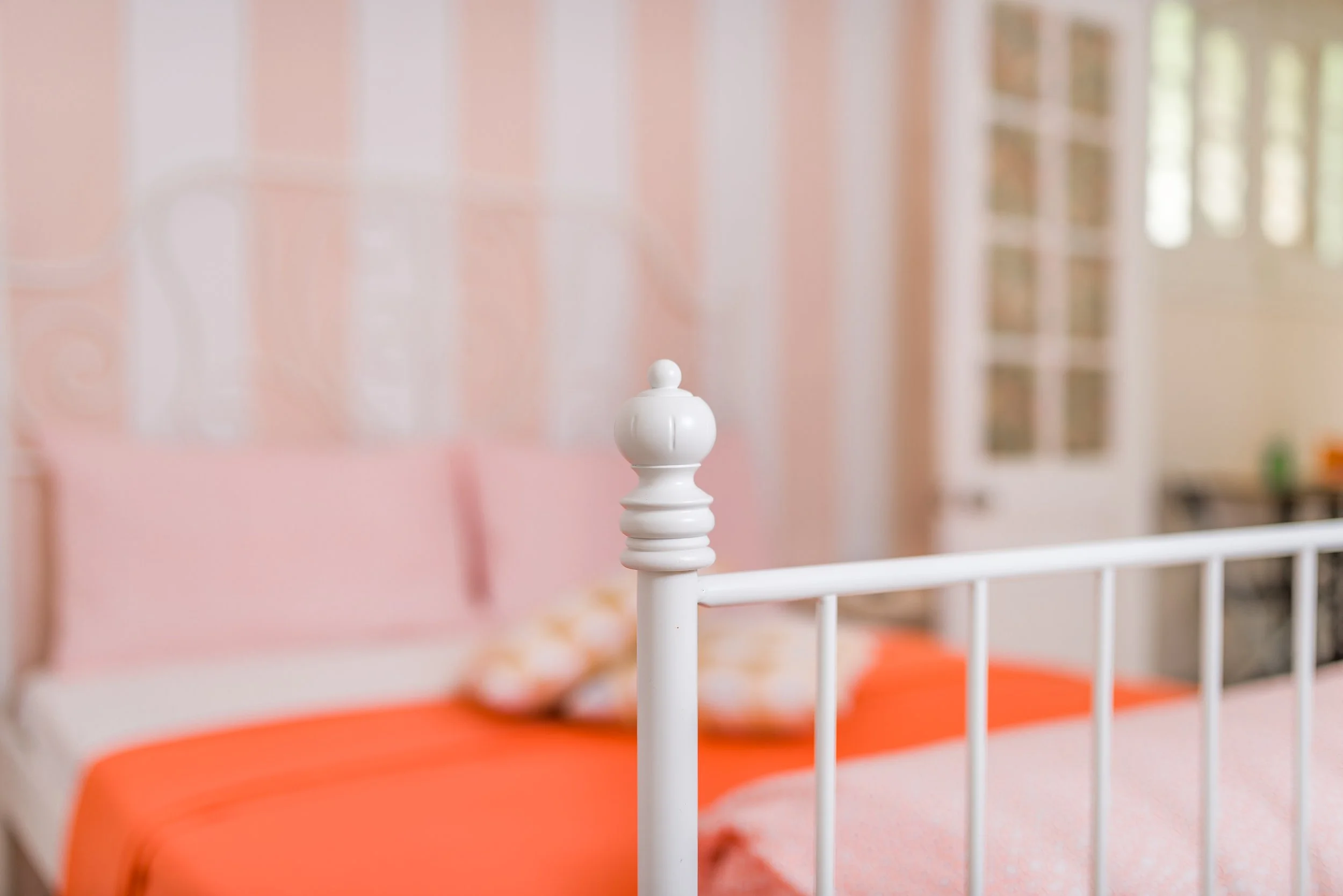 Cerca de la cabecera de una cama, la barandilla blanca de la cama y una colcha de color naranja. En el fondo, la pared rosa y ventanas que permiten la entrada de luz natural al cuarto.