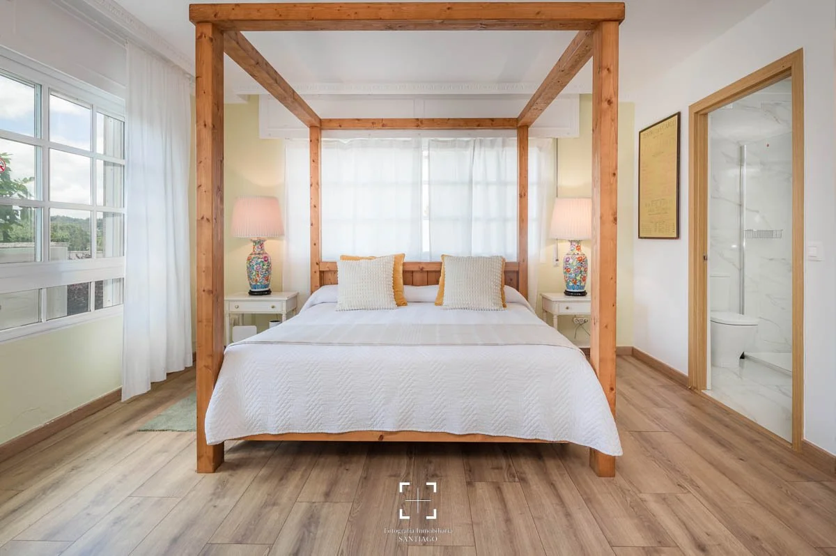 Habitación con cama de madera con dos cabeceros, cortinas blancas, dos lámparas con bases decorativas, y baño visible a la derecha.