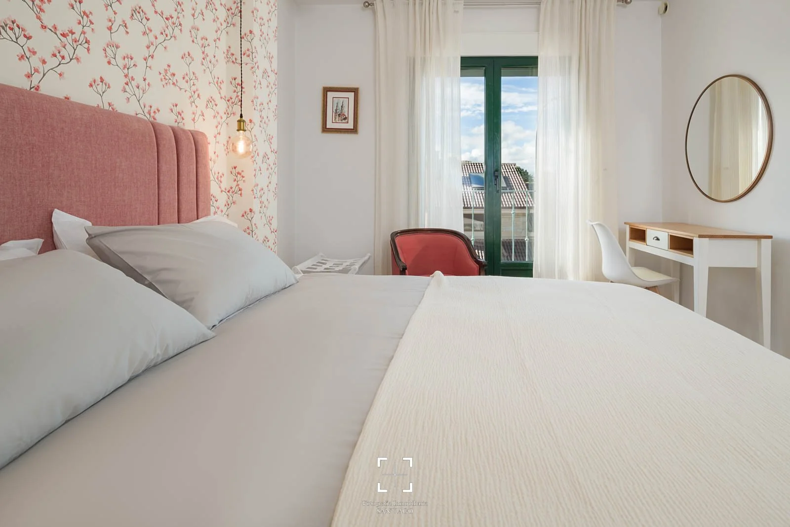 Habitación con paredes decoradas con papel tapiz de ramas con flores rosas, cama con cabecera rosa, pillows blancos, lámpara de luz cálida colgante, ventana con cortinas blancas, vista a tejados y cielo, escritorio pequeño con espejo redondo, sillín 