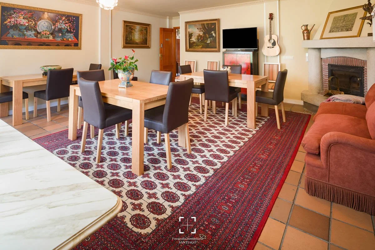 Sala de comedor con cuatro mesas de madera y sillas tapizadas en cuero negro, una alfombra de diseño en tonos rojos y beige, una chimenea, un sofá antiguo en tono terracota, paredes con cuadros y pinturas, una guitarra colgada en la pared, y ventanal