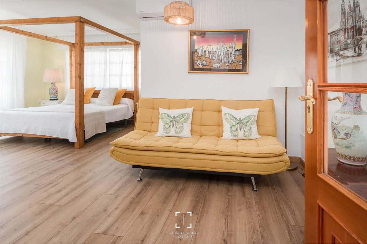Sala con sillón amarillo y cojines con mariposas, habitación con cama con dos almohadas naranjas y blancas, lámpara de pie blanca y lámpara de mesa con base decorativa, cuadro en la pared, y puerta de madera parcialmente abierta, decorada con un jarr