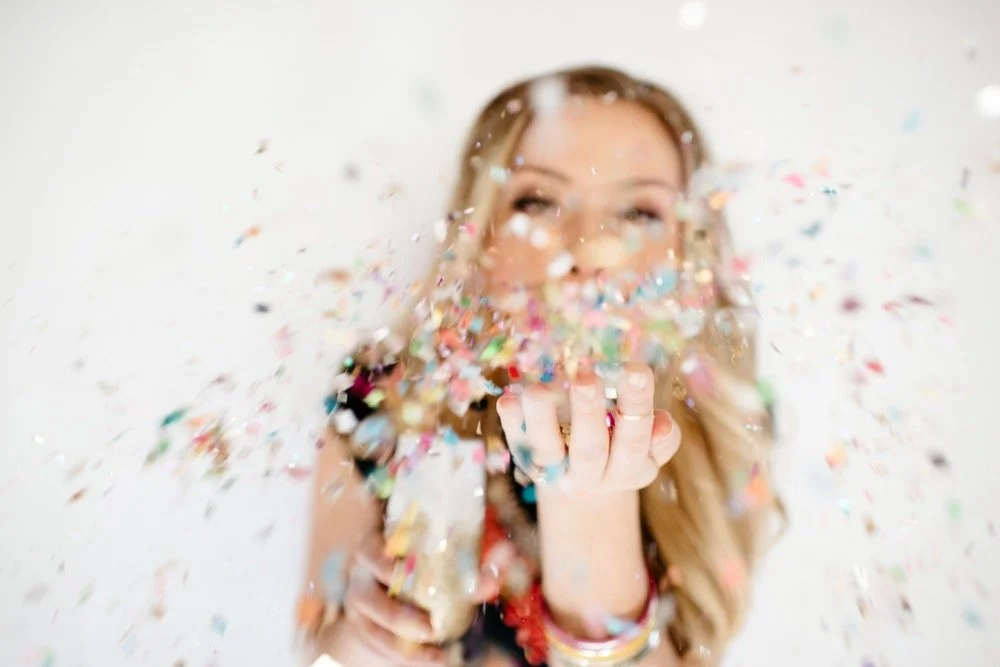 blonde haired girl blowing a handful of colorful confetti so it's exploding toward the viewer