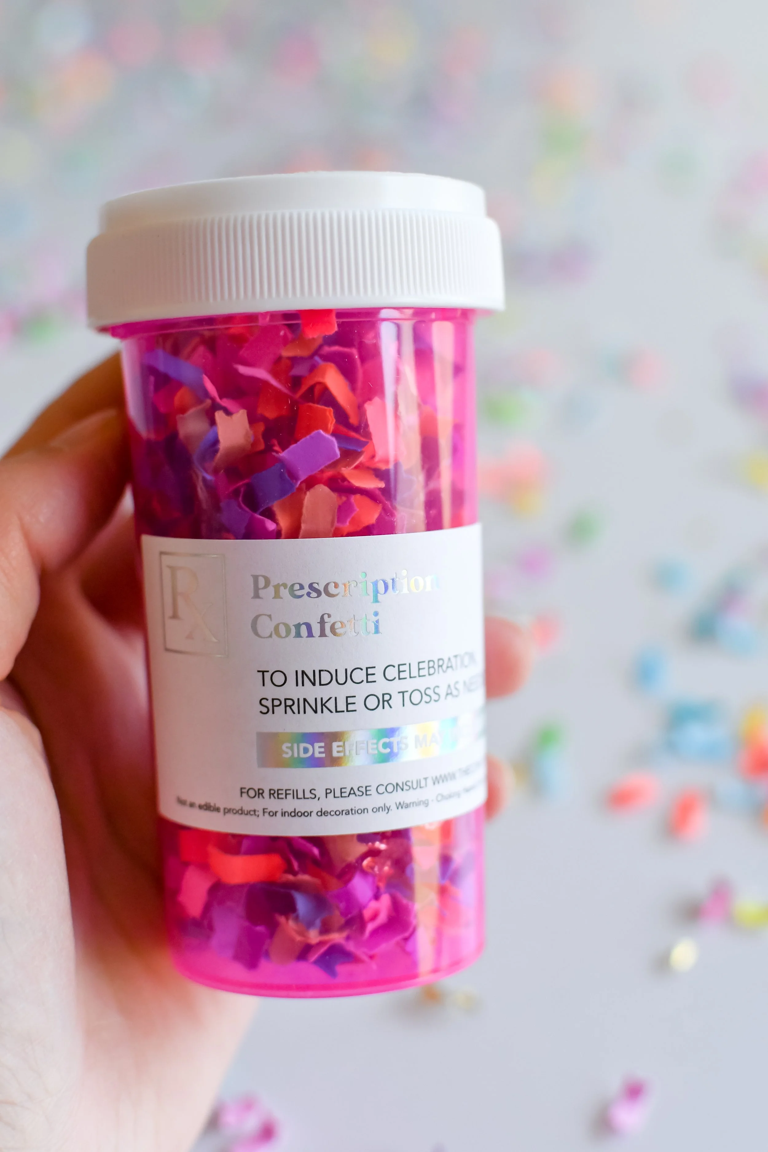 A hand holding a pink container of colorful confetti with a label that reads 'Prescription Confetti,' used for celebration, sprinkled on a surface with more confetti scattered around.