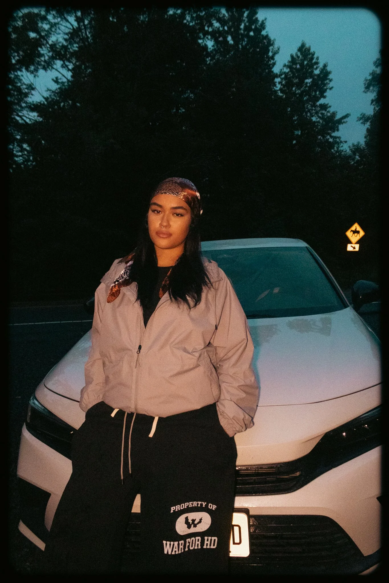 A woman standing in front of a white car at dusk. She is wearing a beige jacket, black pants with text, and a bandana on her head. There is a wildlife crossing sign in the background and trees are visible in the sky.