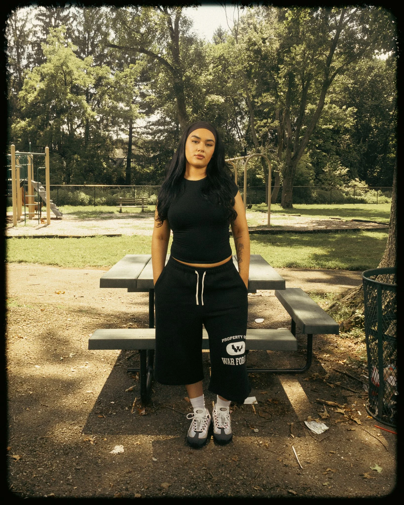 A young woman with long black hair, wearing a black crop top, black pants with white text, white socks, and sneakers, standing outside in a park near a picnic table, with trees and playground equipment in the background.