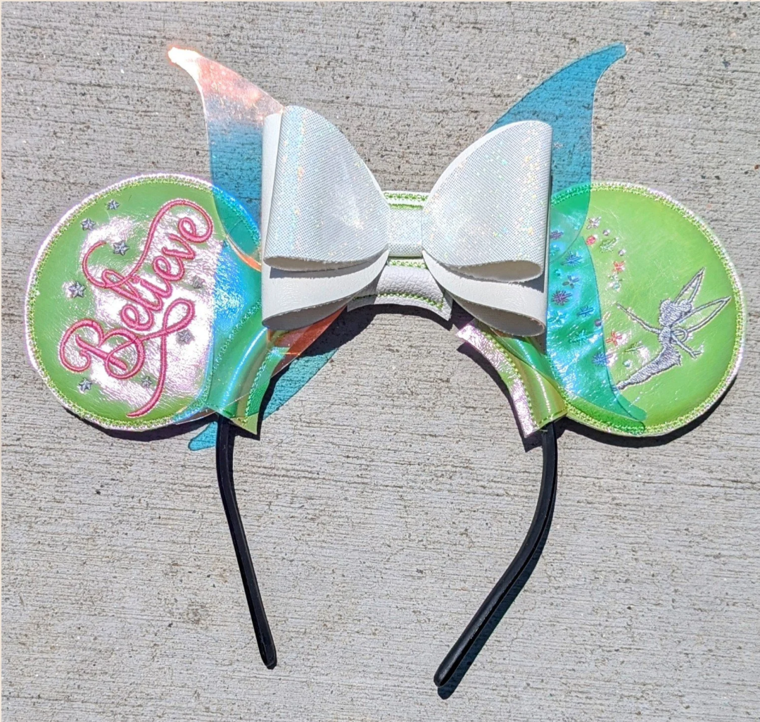 A flexible headband featuring Tinker Bell, with a large white bow, holographic ears reading 'Believe', and blue and green holographic pixie wings, laid on a concrete surface.