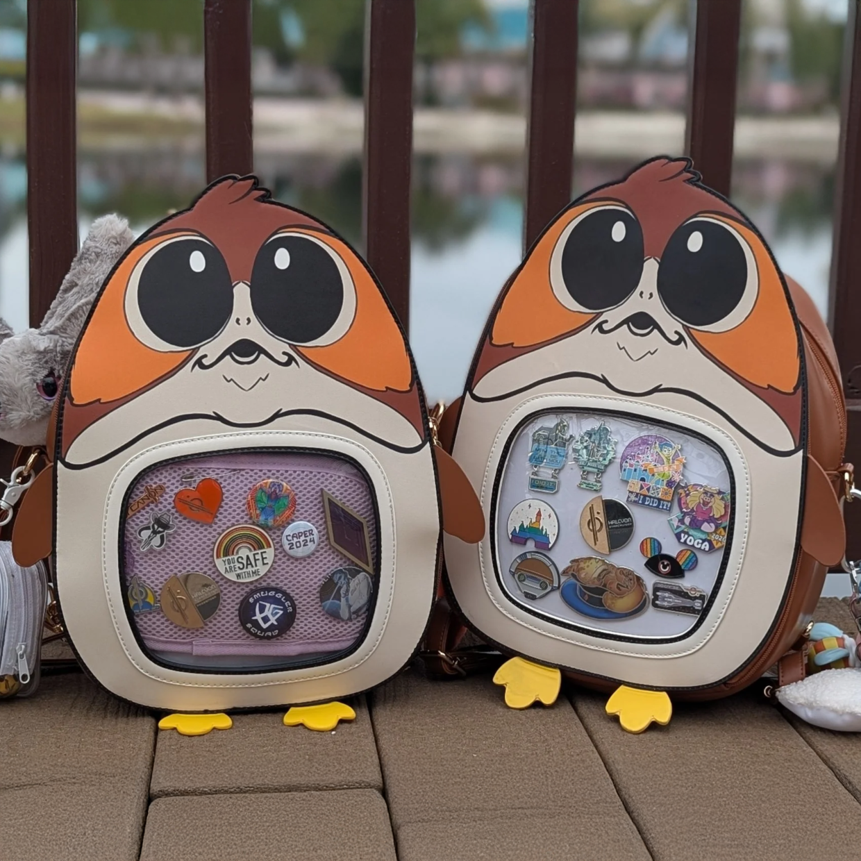 Two backpacks designed to look like  porgs, filled with colorful pins and placed on a wooden boardwalk outdoors near a body of water.