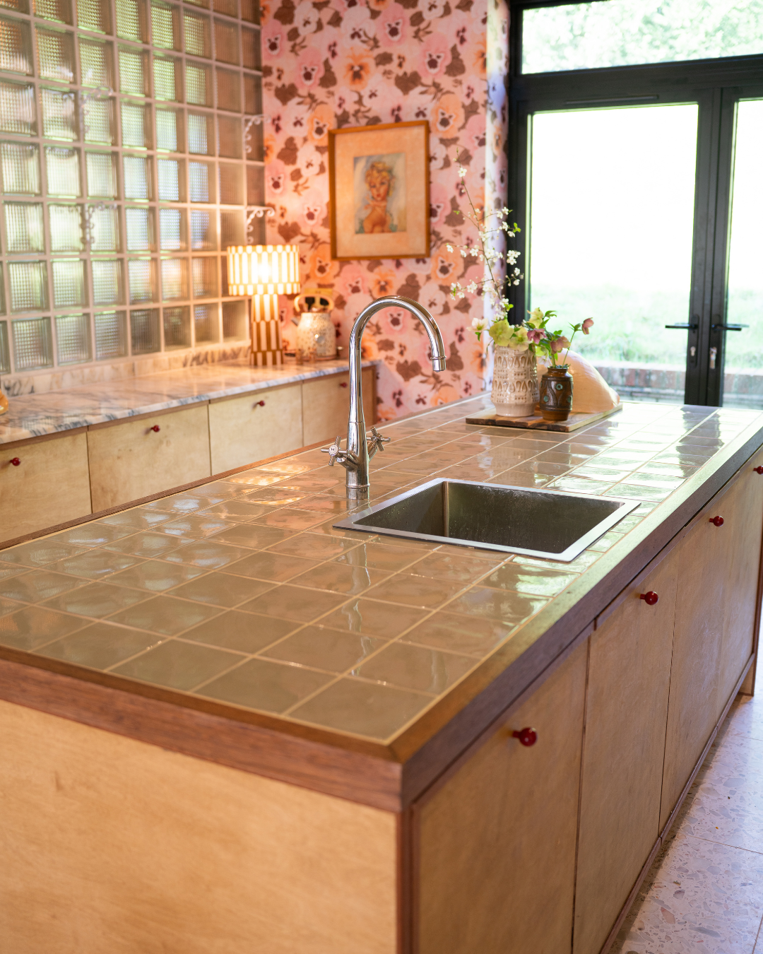 A kitchen with a tiled countertop, a sink with a chrome faucet, decorative vases with flowers, a lamp with a striped shade, floral wallpaper, a framed picture of a woman, and glass doors leading outside.