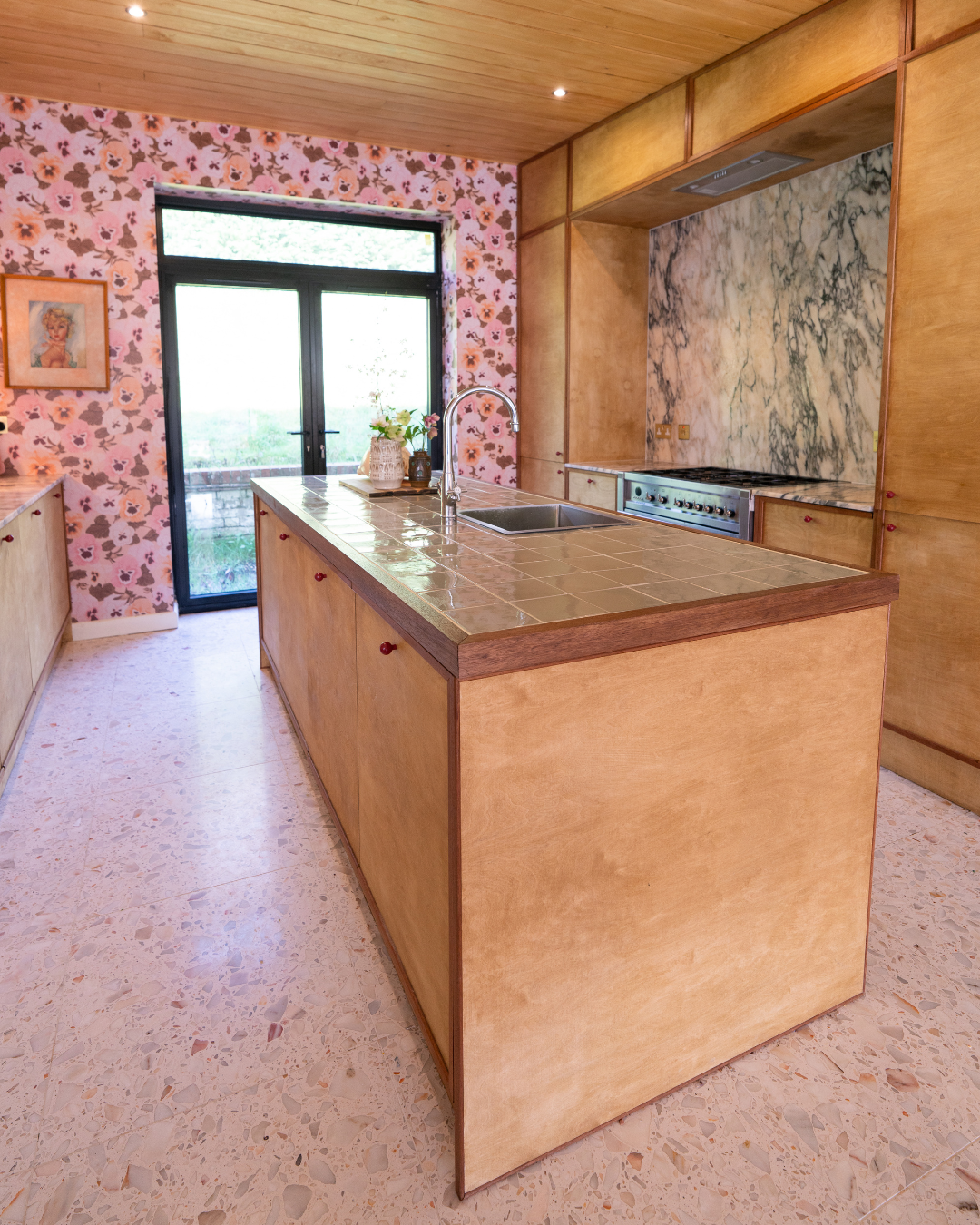 Kitchen island with beige tile top, wooden base, and a faucet, next to sliding glass door with floral wallpaper and a picture on the wall.