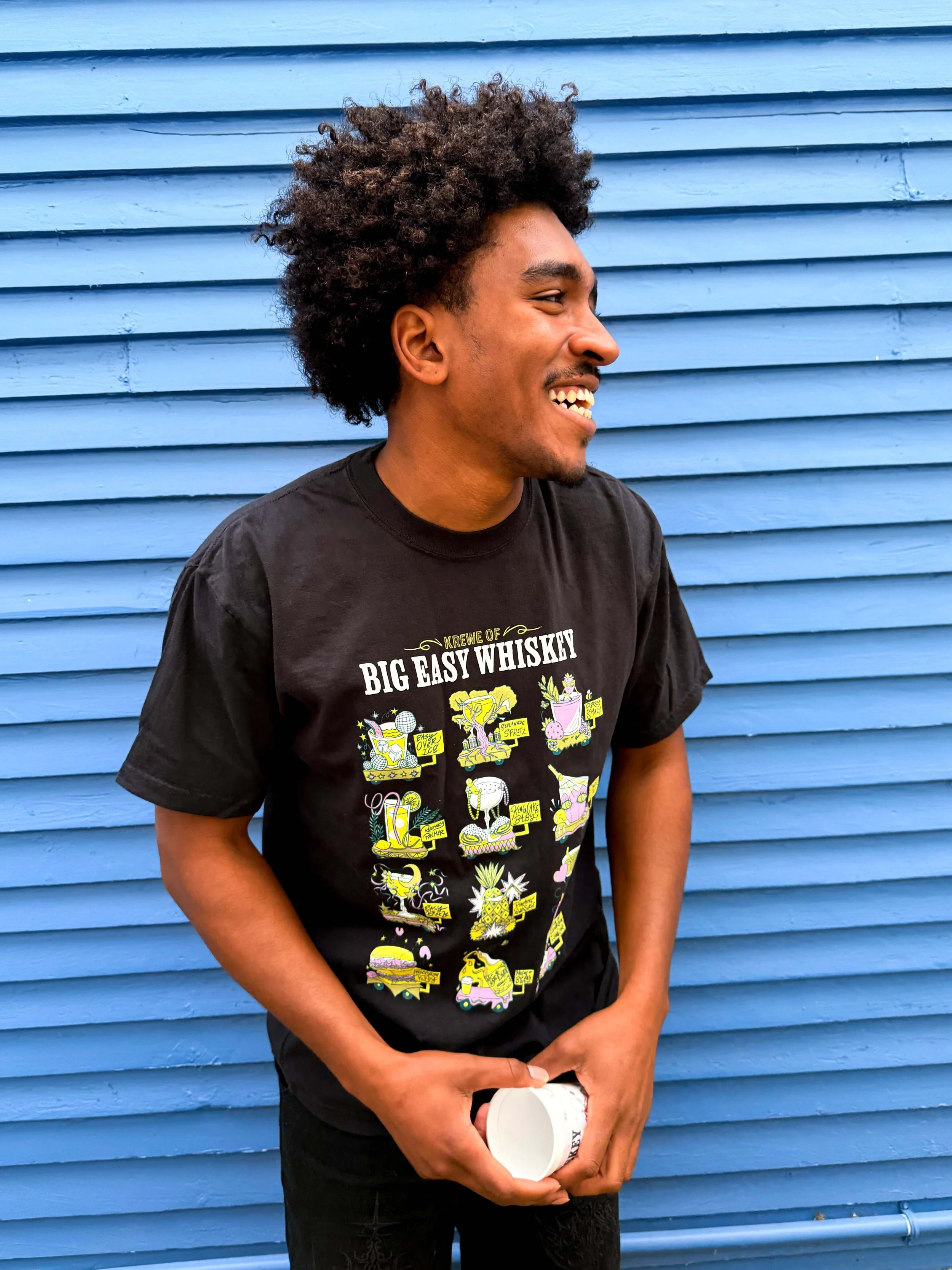 A young man with curly hair smiling, standing against a blue wooden wall, wearing a black t-shirt with colorful graphics and text related to whiskey.