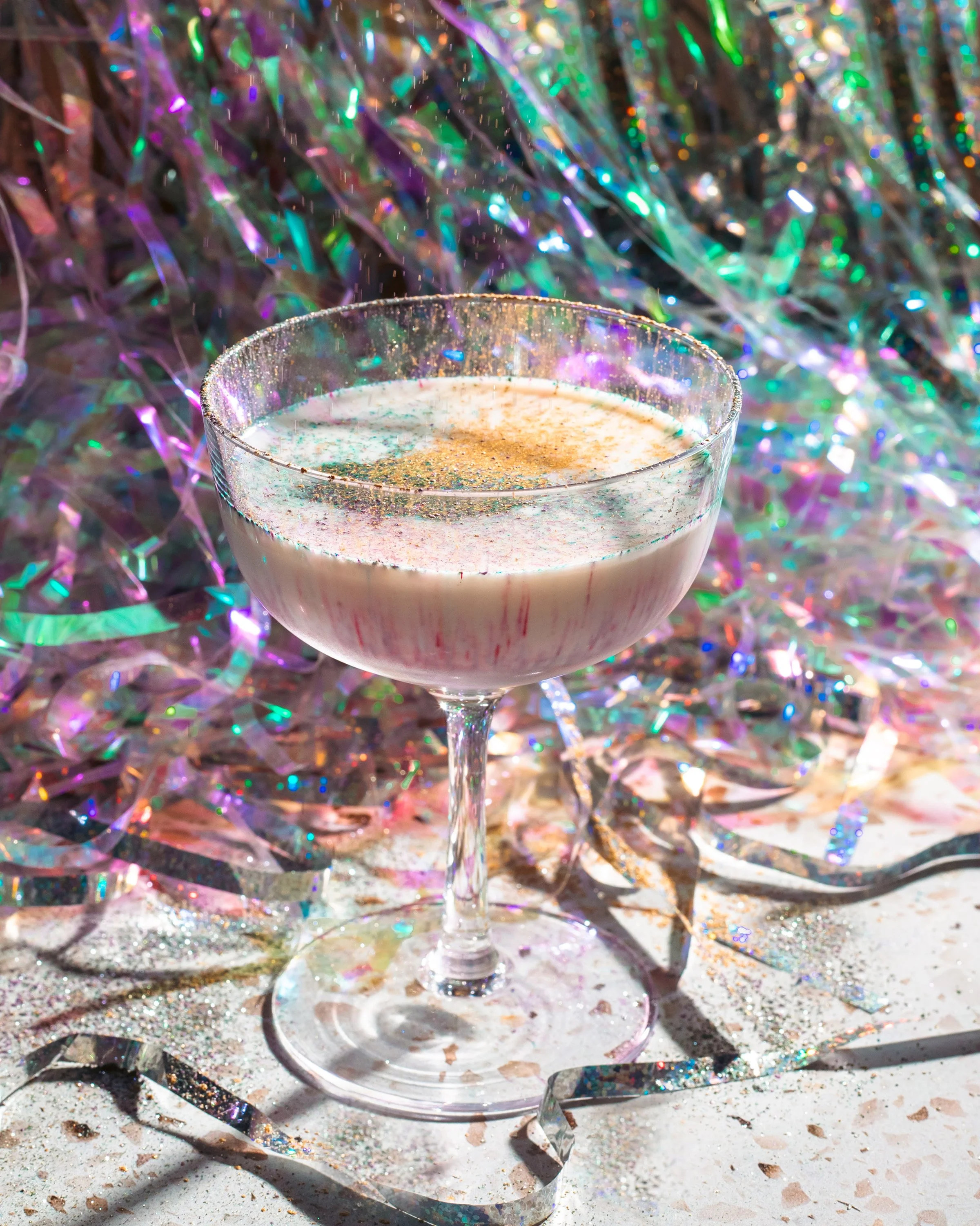 A festive drink in a coupe glass with iridescent confetti and streamers in the background.