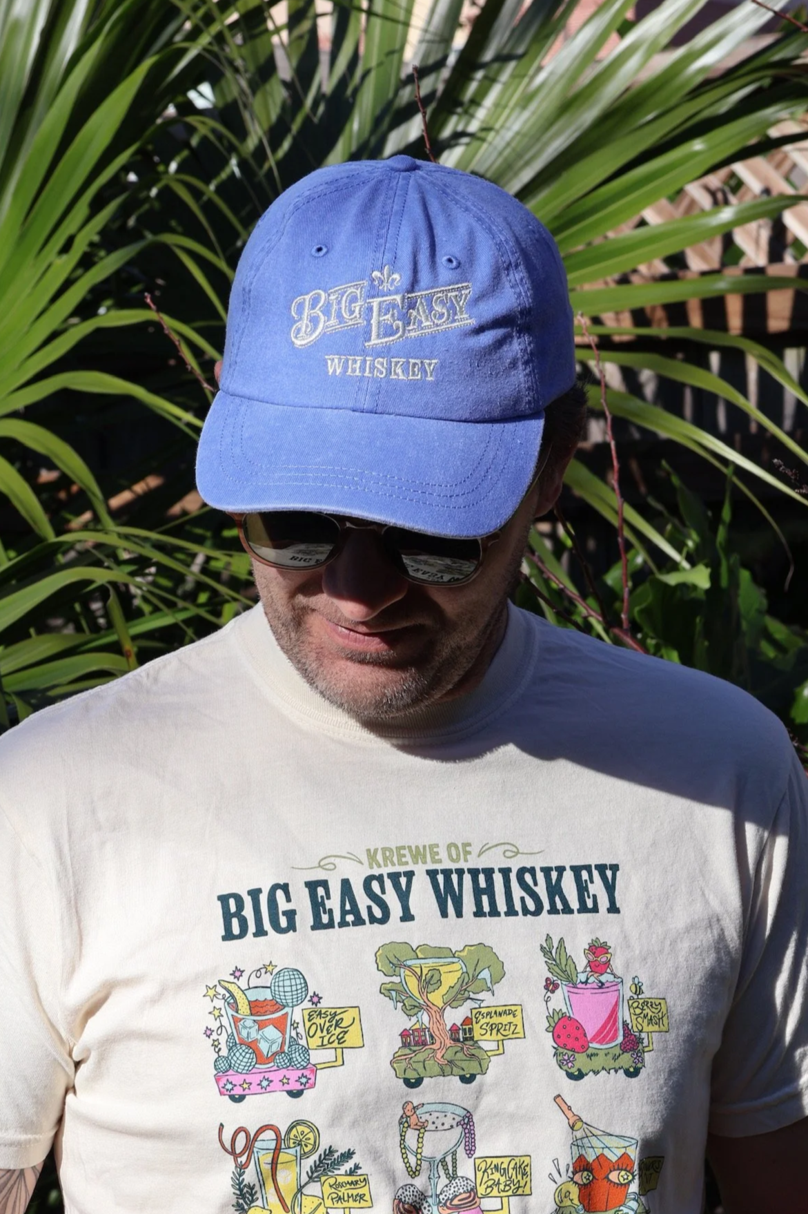 A man is wearing a light blue cap with white embroidered text that reads "Big Easy" and "Whiskey," as well as mirrored sunglasses, and a white T-shirt with colorful illustrations and text related to a Louisiana-themed gathering or event.