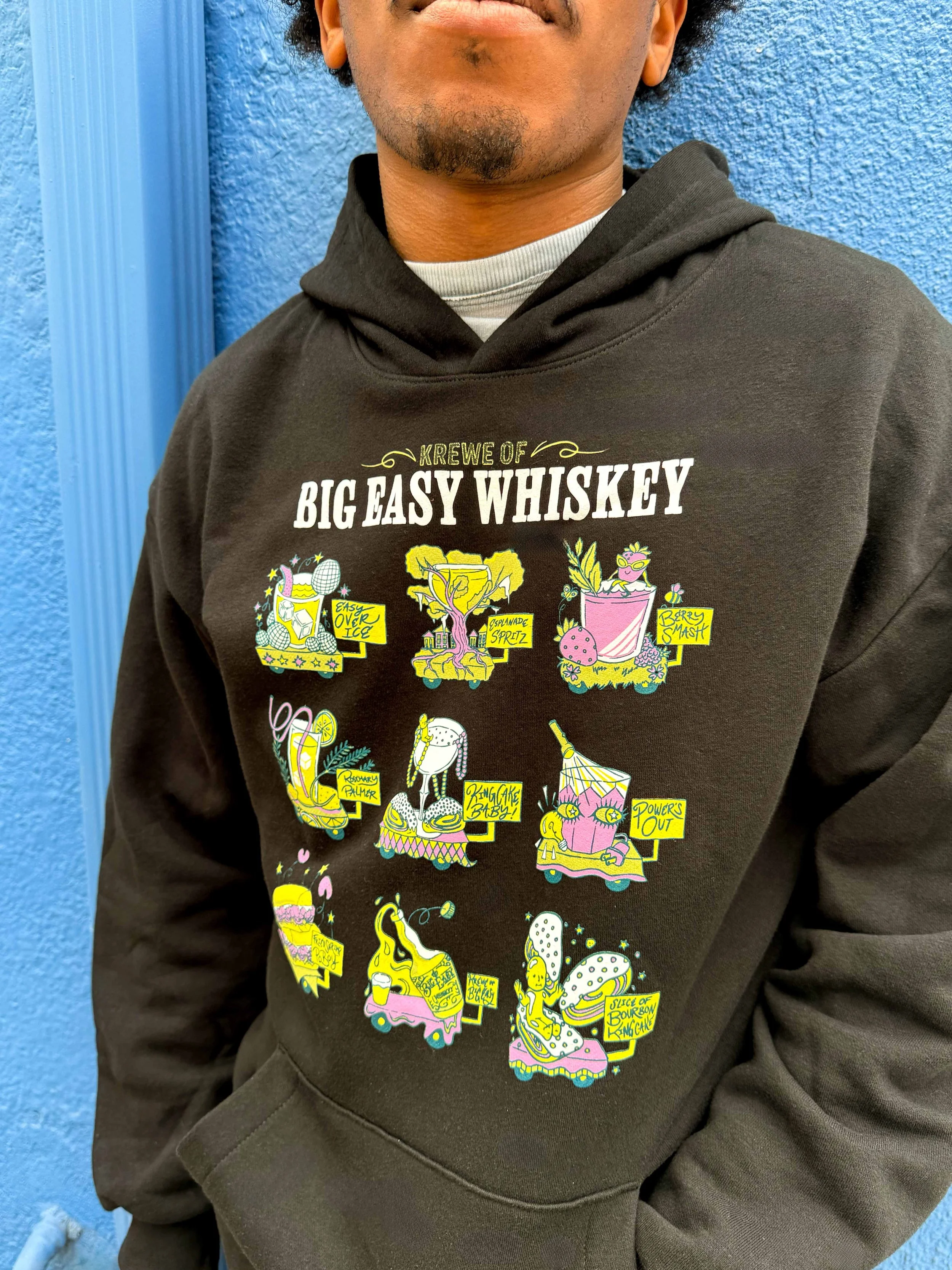 A person wearing a black hoodie with colored cartoon-style illustrations and text related to a humorous theme about a whiskey crew, standing against a blue textured wall.