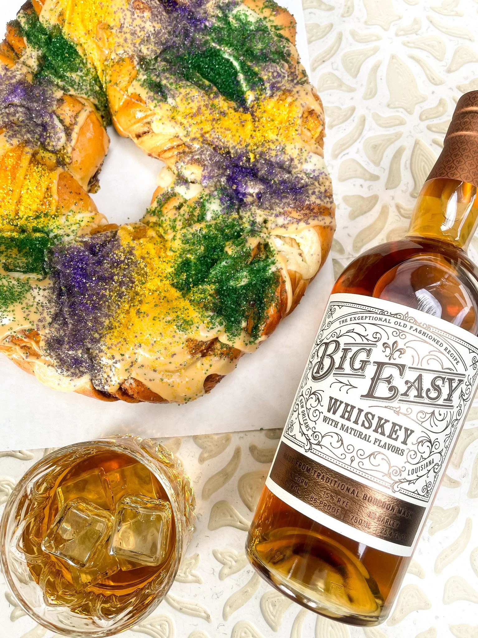 A Mardi Gras king cake decorated with purple, green, and yellow sugar, a glass of whiskey with ice, and a bottle of Big Easy whiskey on a patterned table.